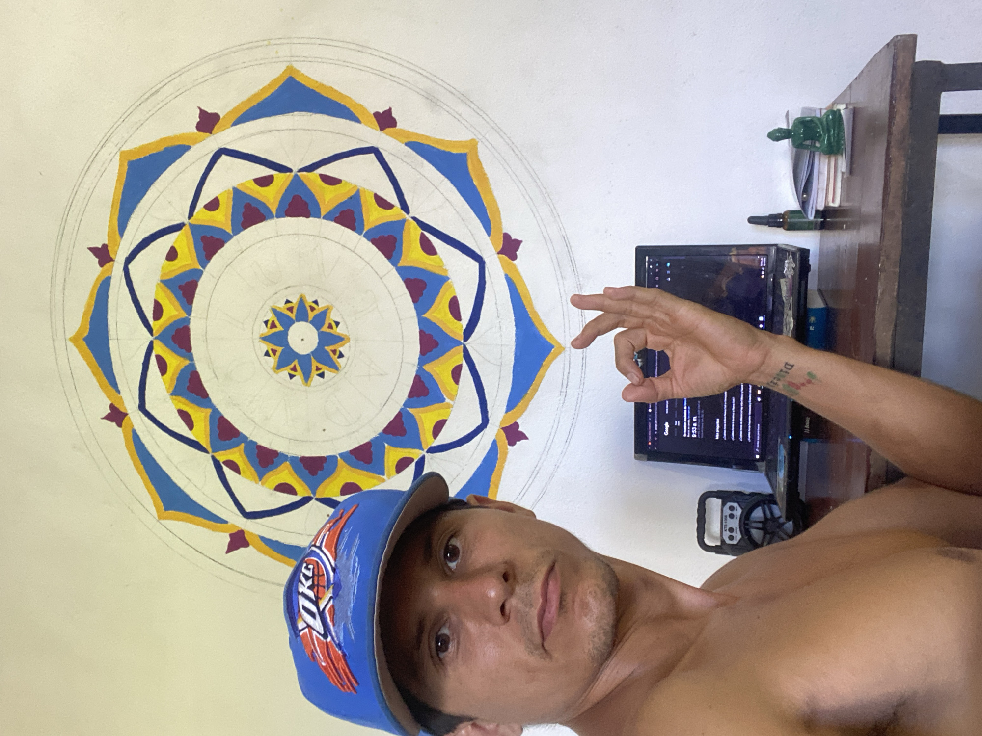 A man is posing for a selfie in an indoor environment. He is wearing a blue baseball cap with the logo "OKC" on it. He is shirtless and has fair skin. His right hand is raised, forming a circle with his thumb and index finger. A tattoo is visible on his left forearm.
Behind him on the wall is a large, colorful mandala painting. The design features concentric circles with geometric patterns and various colors including yellow, blue, and purple.
To the right of the man, there's a wooden desk with a computer monitor on top of it. A black speaker is positioned below the monitor. There's also a green figurine or object, along with some papers, stacked on top of the desk near the edge.
The location is identified as Los Velásquez, Venezuela.