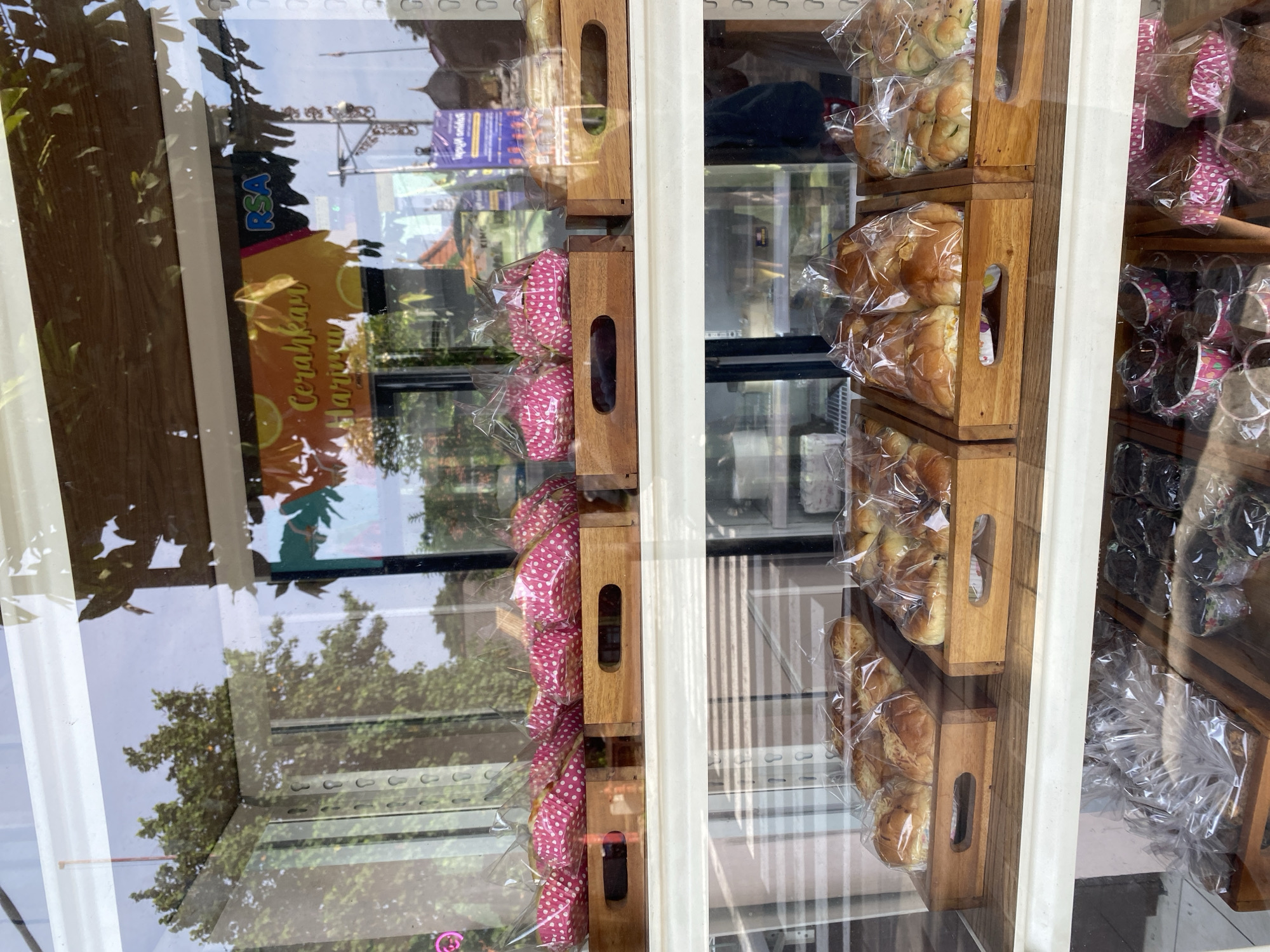 A display window in Nusa Dua, Indonesia shows baked goods arranged in wooden crates. The items include breads and cakes in polka-dotted wrappers, all sealed in plastic bags. The window reflects the surroundings, including trees and signage. A commercial refrigerator can be seen behind the baked goods.