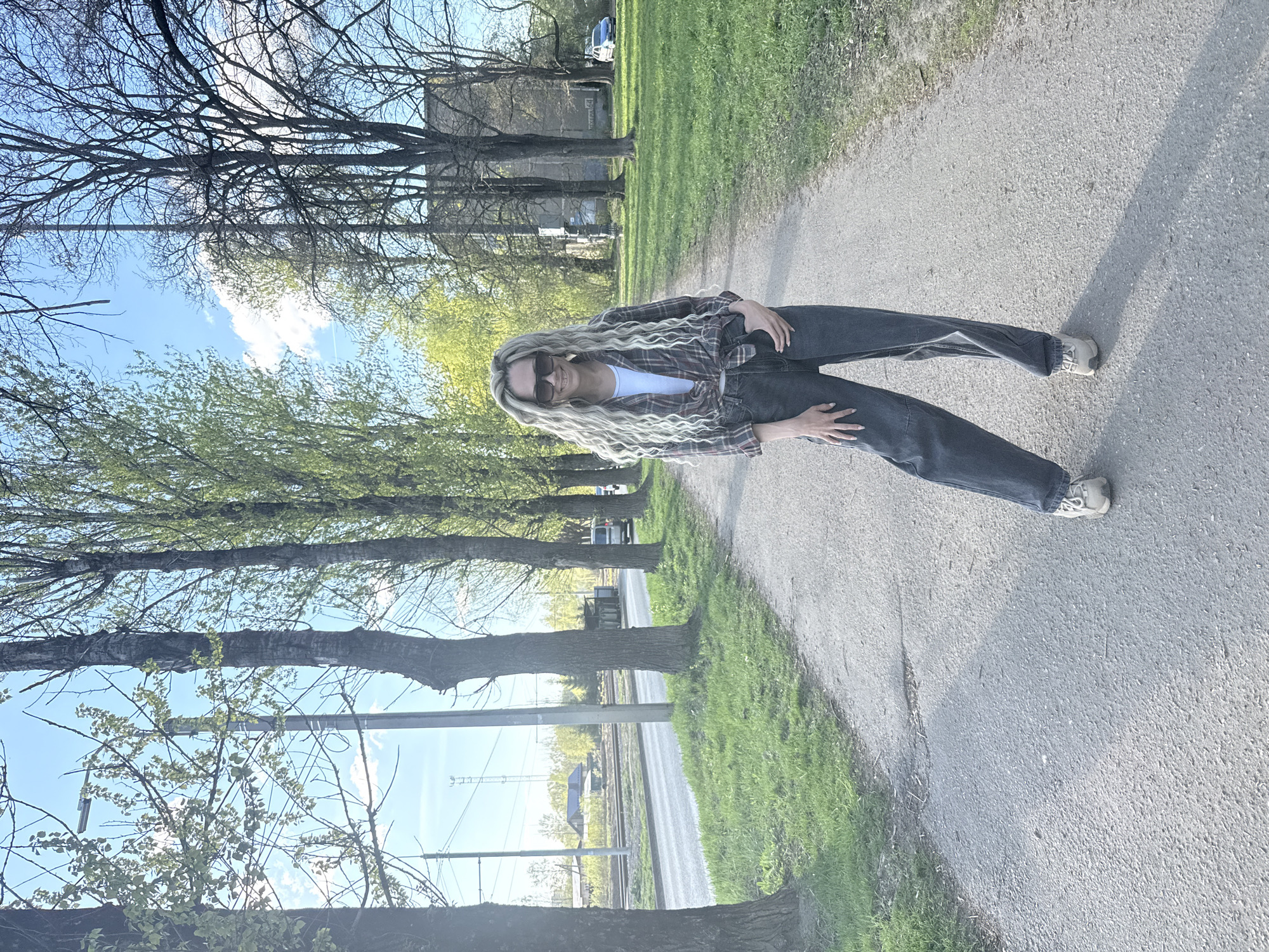 A woman stands on a paved path, seemingly in a park or green area. She wears sunglasses, a plaid shirt tied at the waist over a white top, dark jeans, and light-colored sneakers. She has long, light blonde hair. The path has grass on either side and is lined with trees. In the background, there are buildings, including what looks like a railway infrastructure. This image appears to be shot near Rusanovo, Russia, given the user's prompt. The location would be near train tracks.