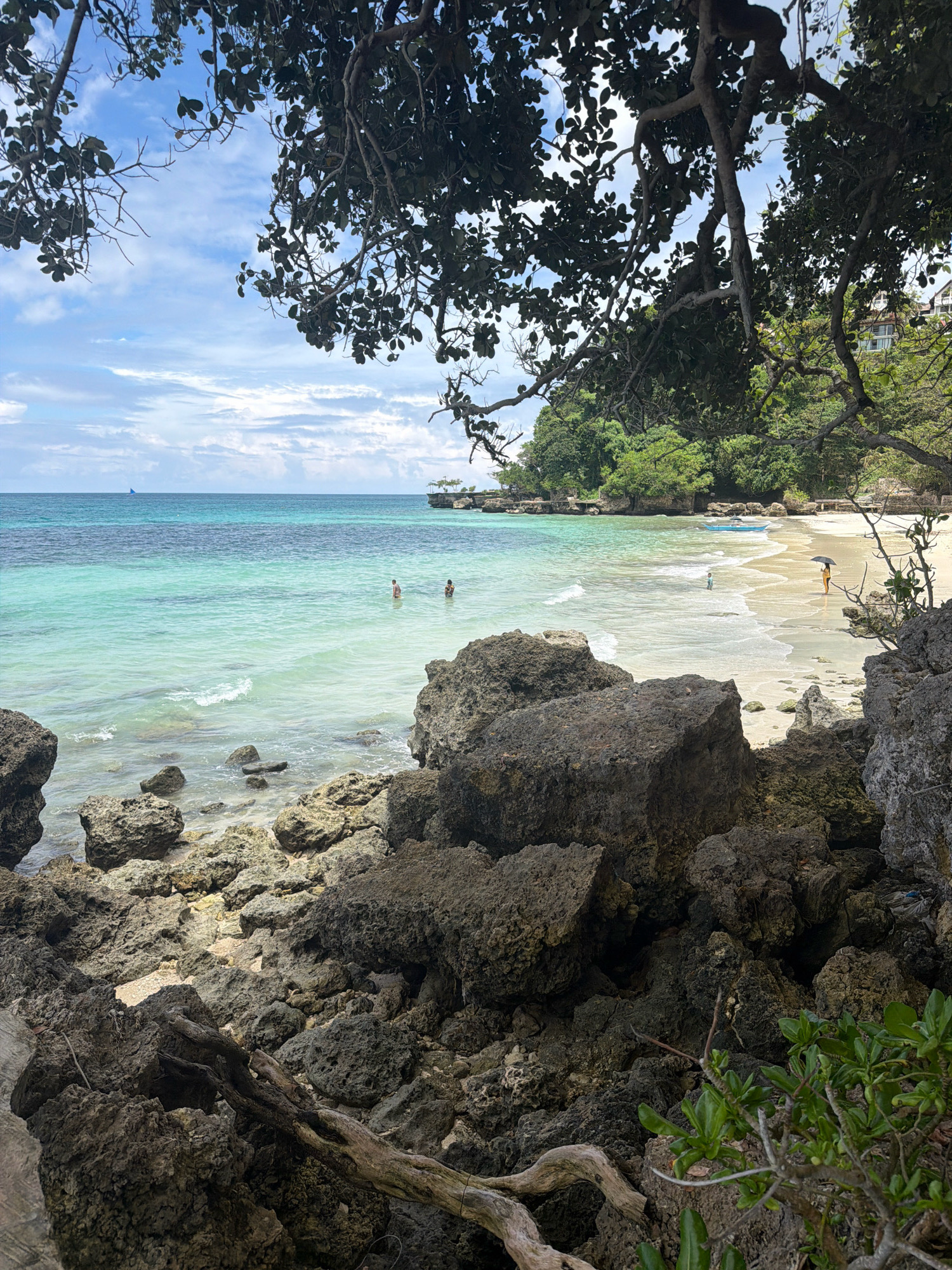 The image depicts a tropical beach scene, likely in Malay, Philippines. The foreground is dominated by dark, rugged volcanic rocks and driftwood, partially obscuring the view of the beach and sea. Beyond the rocky shore, the turquoise waters of the ocean are clear, with gentle waves lapping at the sandy beach. In the shallow water, two individuals are visible, appearing to be swimming or wading. Further along the beach, a person holds a black umbrella, standing near the water's edge. The background features lush green vegetation and trees, with buildings peeking through the foliage, suggesting a resort or residential area. The sky is partly cloudy, with a serene blue expanse. A small sailboat is visible on the distant horizon.