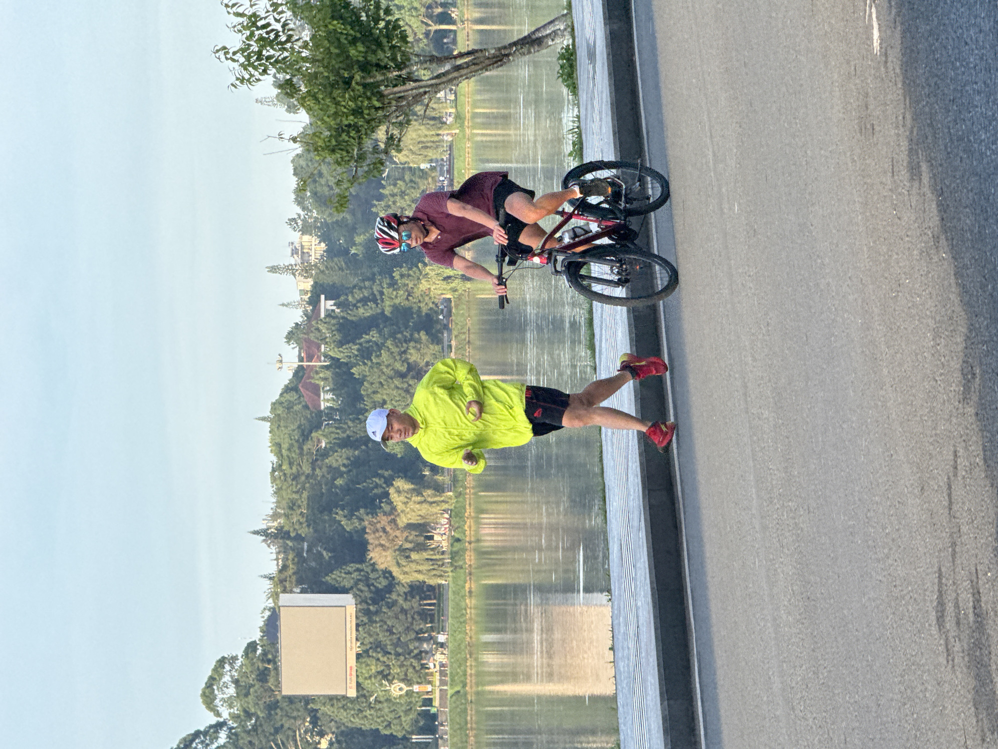 The image captures a waterfront scene in Da Lat, Vietnam, likely along Xuan Huong Lake, during the early morning or late afternoon given the soft lighting. A man in a bright yellow jacket and black shorts is jogging on a paved pathway beside a body of water. He is wearing a white cap and red athletic shoes. To his right and slightly behind him, another man is riding a bicycle on the same pathway. This cyclist is wearing a maroon polo shirt, black shorts, a red and white helmet, and sunglasses. His bicycle is dark red and black. The pathway is bordered by a low concrete wall with metal grating on top. Beyond the pathway, the calm water of the lake reflects the trees and buildings on the opposite shore. The background features a dense line of trees, some buildings, and a large, light-colored rectangular billboard or screen. The overall atmosphere is one of healthy outdoor activity in a picturesque urban setting.
