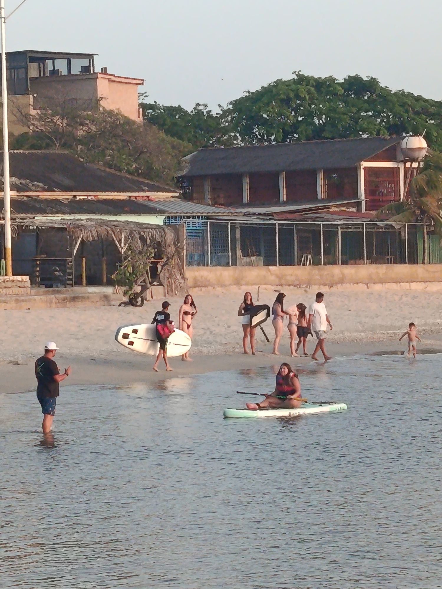 The image depicts a beach scene in Juan Griego, Venezuela. Several people are present, including individuals with surfboards, a group standing near the shoreline, a child playing in the water, and a woman paddling on a stand-up paddleboard in the foreground. A man stands in the water near the shoreline, appearing to observe or photograph the scene. In the background are various buildings, including structures with wooden and thatched elements, and a building with a water tank on top. Lush green trees are visible further in the distance. The sky is clear and bright, suggesting daytime.