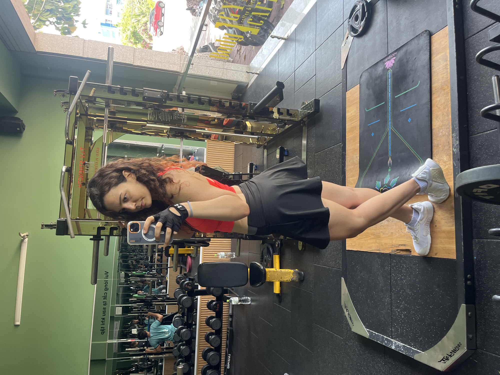 A woman is taking a selfie in a gym located in the city of Phan Thiet, Vietnam. The woman is wearing a red sports bra, a black skirt, and white sneakers. She has curly brown hair and is wearing black workout gloves. The gym is equipped with various weightlifting machines and dumbbells. In the background, there are other gym-goers working out. The gym has green walls with reflective surfaces and what appears to be a large mirror. Through the glass windows of the gym, a street scene is visible with a red car and other buildings. The overall environment suggests a well-equipped fitness center.