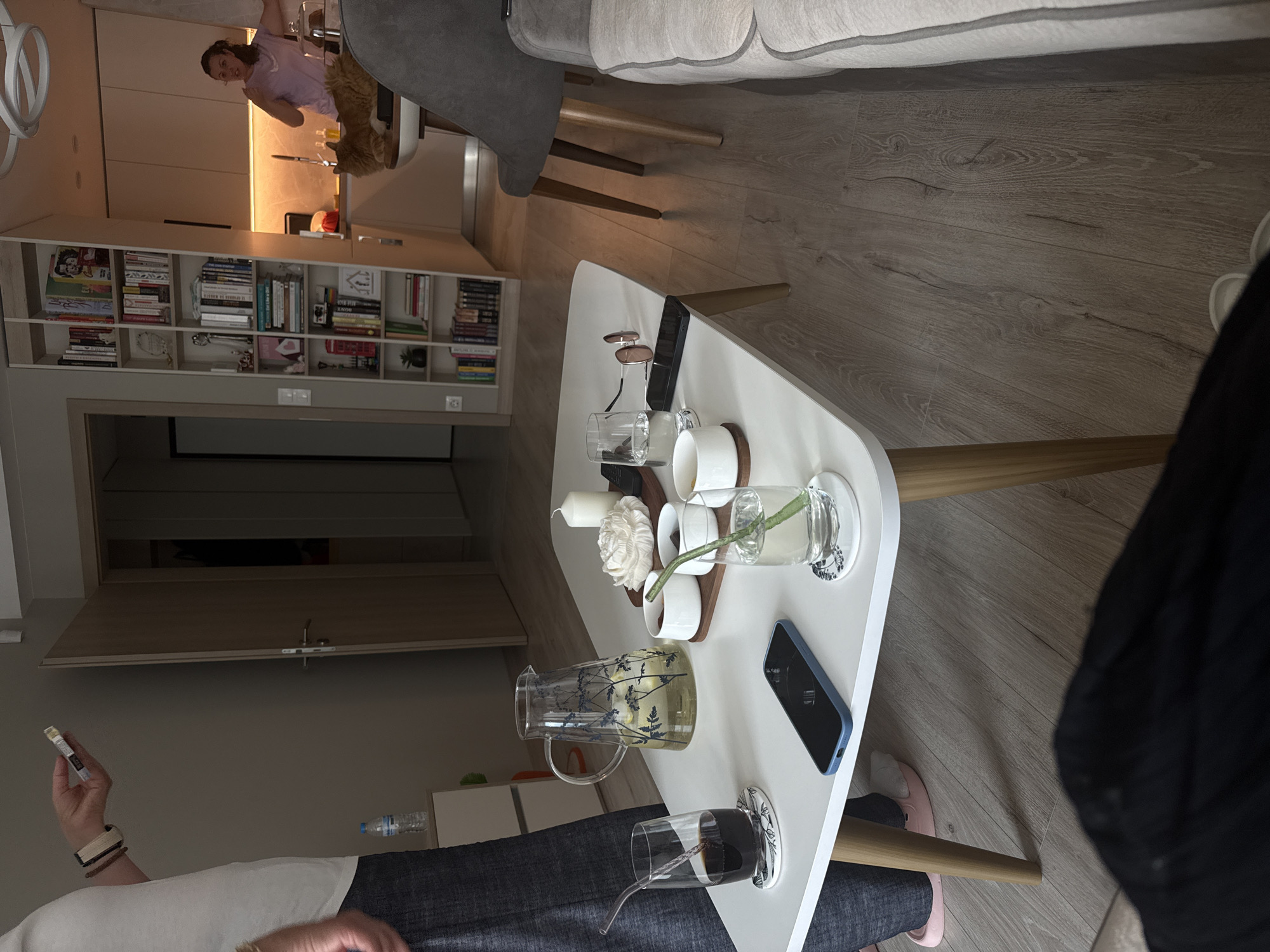 The image depicts an indoor scene within a modern apartment located in Sofia, Bulgaria, showcasing an open-concept living room and kitchen area.
In the foreground, a white rectangular coffee table with four light wooden legs occupies the central space. On the table, from left to right, are: a clear glass containing a dark beverage with a glass straw, resting on a white coaster with a blue floral design; a blue-cased smartphone placed face down; a large glass pitcher with a blue botanical design, containing water with lemon slices; a white ceramic candle; a white decorative object resembling a large rose; a small wooden tray holding four small white ceramic bowls; a black smartphone; sunglasses with round brown lenses and a gold frame; and a black remote control. To the right of these items, a clear glass contains water with a green straw, resting on another white coaster with a blue floral design.
To the left of the coffee table, a person is partially visible, standing. This individual wears a white short-sleeved t-shirt, dark trousers or jeans, and pink slip-on shoes. Their left forearm is visible, wearing two wristbands (one beige, one dark), and their hand holds a white and gold pack of Marlboro Gold cigarettes. A plastic bottle of water rests on a small white cabinet behind them.
In the mid-ground, a light wood laminate floor extends throughout the visible area. A tall, narrow bookshelf, also in a light wood finish, is positioned vertically, separating the living area from the kitchen. It is filled with books and various decorative items. To the right, a grey upholstered chair with wooden legs is partially visible. The extreme foreground right shows a portion of a light-colored sofa, implying the viewer's perspective.
In the background, the kitchen area features light-colored cabinets with under-cabinet lighting. A woman with dark hair, wearing a light purple crew-neck t-shirt, stands in the kitchen, gesturing with her hands. Her facial expression appears animated. An orange or ginger-colored cat is observed lying on a white counter or table surface in the kitchen, to the left of the woman. A modern white circular light fixture is partially visible on the ceiling in the upper right quadrant. Further in the background, light wooden doors are visible, indicating other rooms or entryways.