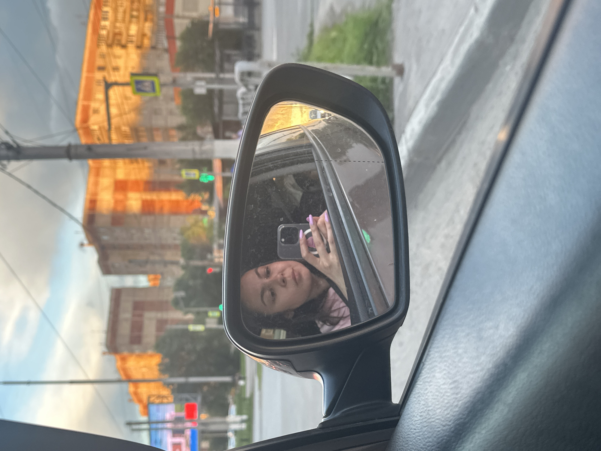 The media file captures a reflection in a car's side mirror, showing a young woman taking a selfie with her phone. She is looking at the camera, with one hand holding the phone and the other, with purple-painted nails, is visible near her face. She has dark hair and is wearing a pink shirt. The reflection also shows the interior of the car, including the window and part of the dashboard.
Outside the car, as reflected in the mirror and visible in the rest of the image, is an urban scene. Tall, multi-story buildings with orange and brick facades dominate the background, suggesting an Eastern European city. A street with a sidewalk and traffic lights is visible. One traffic light displays a green signal. There are also utility poles and wires, common in urban environments. The sky is partly cloudy. The warm, golden light suggests it is either early morning or late afternoon. The overall mood of the selfie is casual and relaxed. The location context provided is Magnitogorsk, Russia.