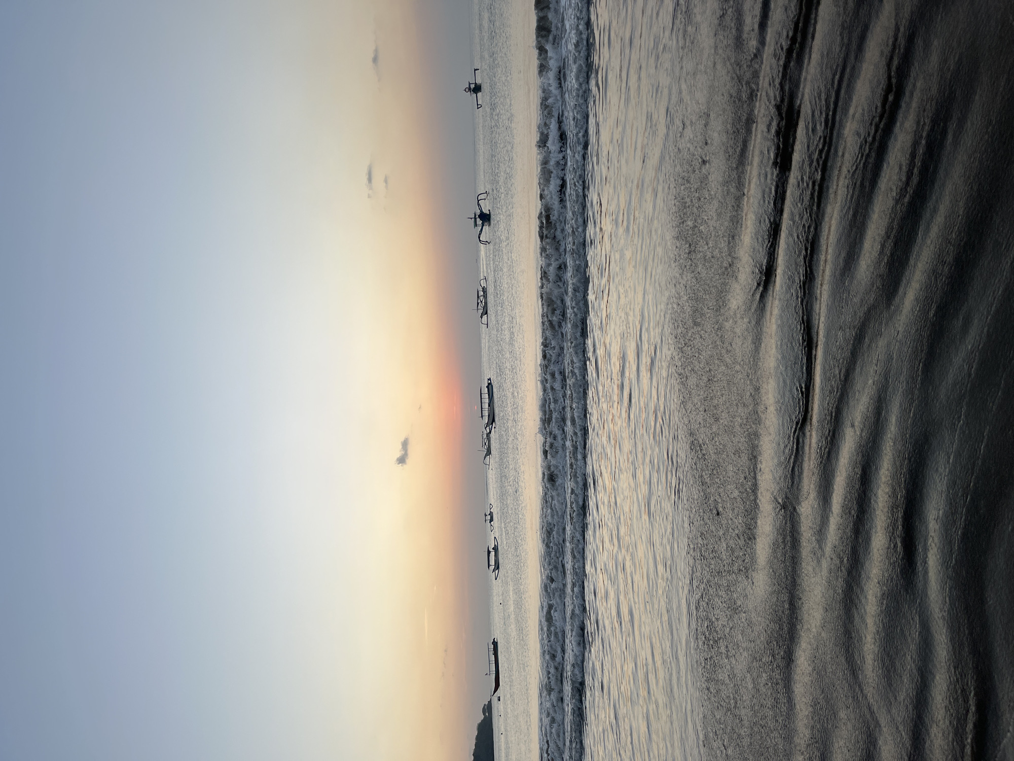 This image depicts a serene beach scene at dusk in Kuta Selatan, Indonesia. The sky transitions from a pale blue at the top to soft hues of orange and pink near the horizon, suggesting either sunrise or sunset. Several traditional Indonesian outrigger boats are visible scattered across the calm ocean, their silhouettes distinct against the water. Gentle waves are breaking near the shore, with the wet sand in the foreground reflecting the muted light and exhibiting ripple patterns. The overall atmosphere is tranquil and peaceful, characteristic of a tropical coastline.