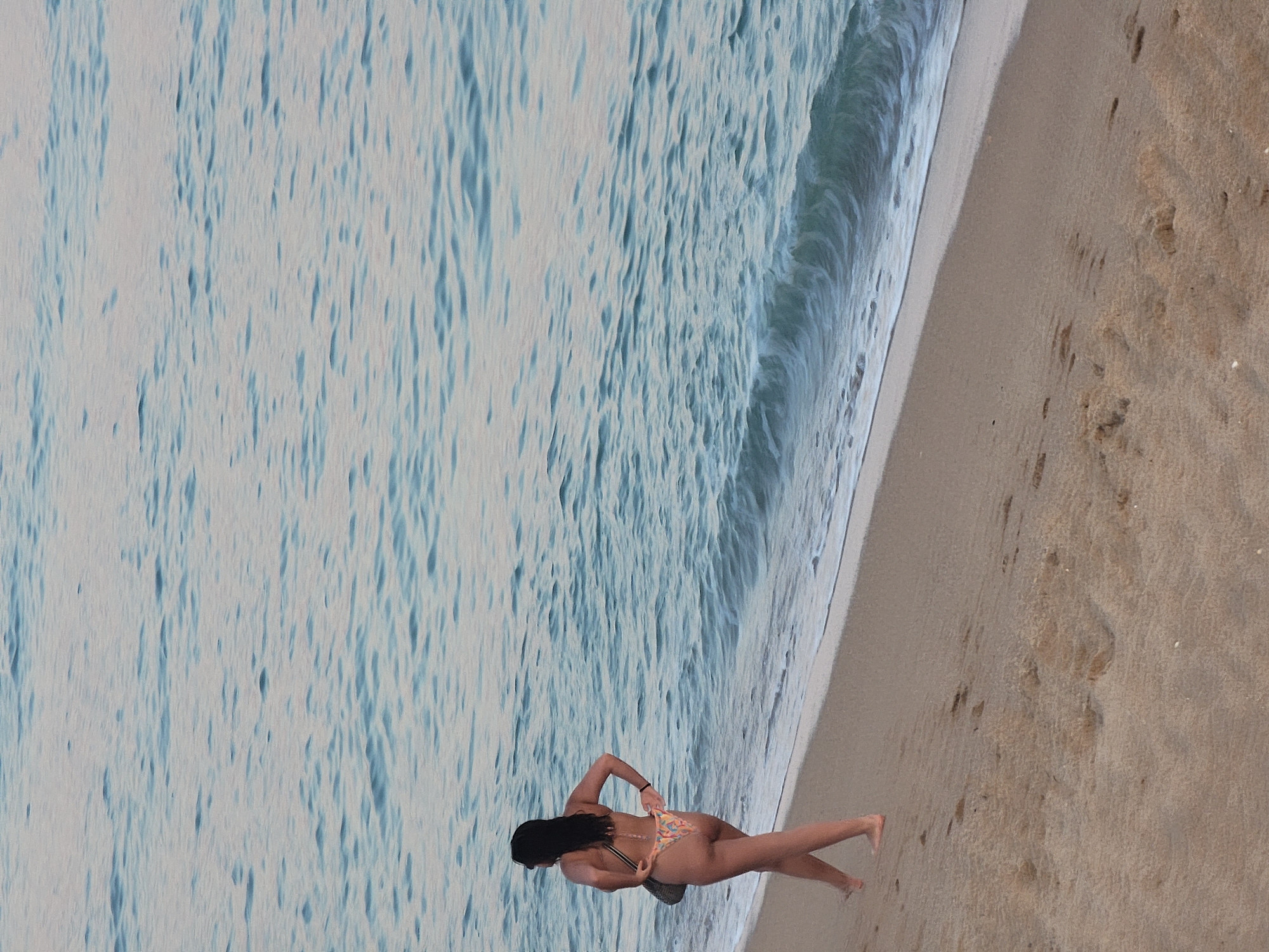 A woman walks along the sandy shore of Quarteira, Portugal, with the gentle waves of the Atlantic Ocean lapping at the beach. She is wearing a bikini and a small bag is slung over her shoulder. The water is a tranquil blue-green, reflecting the soft light of dusk. The sand is damp and imprinted with footprints, suggesting others have walked this path. The scene is serene, capturing a moment of quietude by the sea.