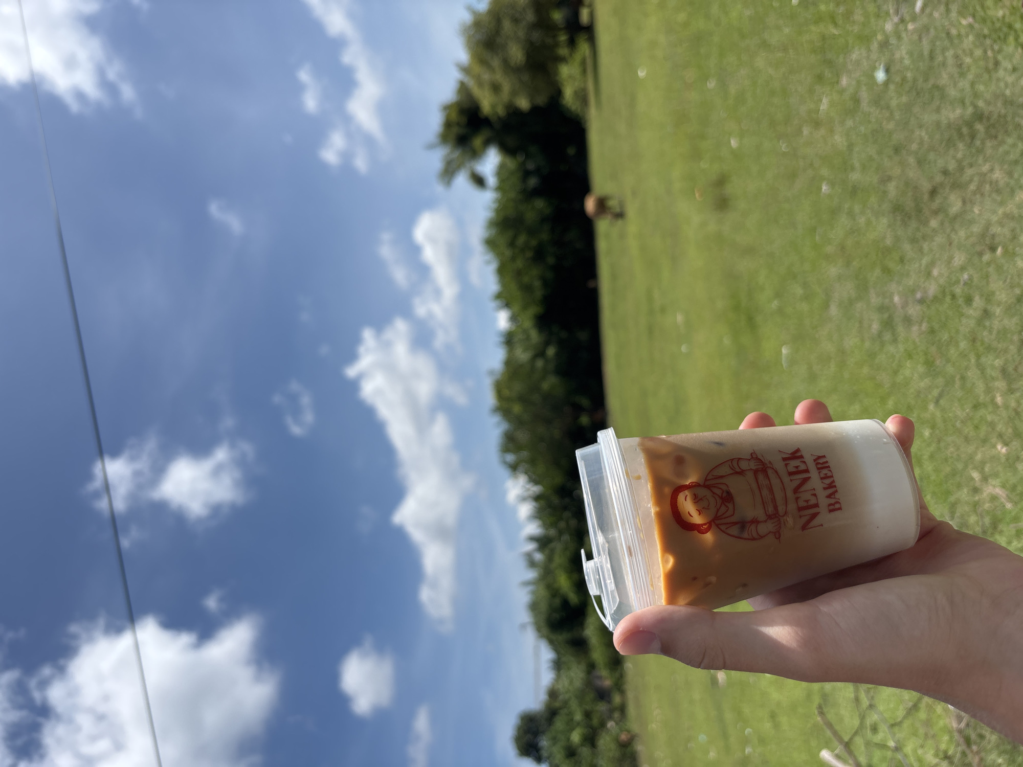 A hand holds a plastic cup containing a layered beverage, presumably coffee, with the label "Nenek Bakery," in an outdoor setting with the sky visible behind it. The background includes a green field and trees, typical of a rural or park-like area. The sky is blue with scattered white clouds. The scene could be in Ungasan, Indonesia, given the bakery name, though the exact location is indistinguishable from this scene.