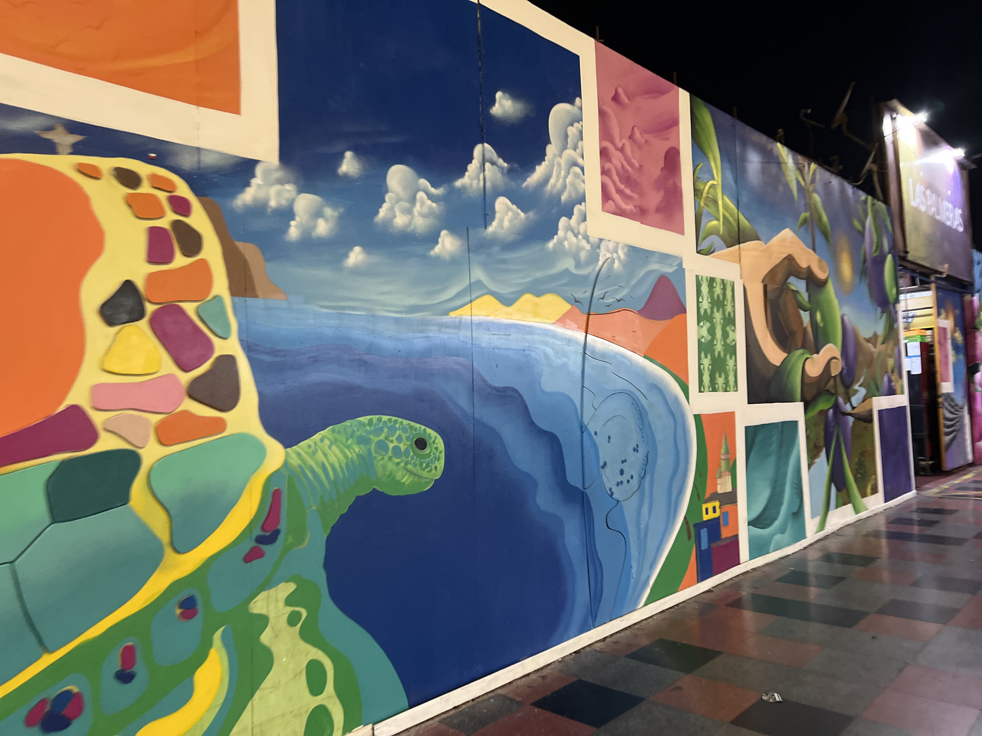 The image depicts a colorful mural painted on a wall in Arica, Chile. The mural features a large sea turtle in the foreground, with its head and flipper visible. The turtle is rendered in shades of green and yellow, with a patterned shell. Behind the turtle, the mural depicts a seascape with a blue ocean, a sandy beach, and rolling hills in various shades of orange, purple, and yellow. The sky is a deep blue with white, fluffy clouds. The mural is sectioned into different panels, each showcasing distinct imagery, including abstract patterns and what appears to be a hand reaching for a plant. In the background, a sign reads "LAS PALMERAS." The mural is illuminated by artificial lighting, suggesting it is nighttime. The ground in front of the mural is paved with checkered tiles in shades of gray, brown, and red.