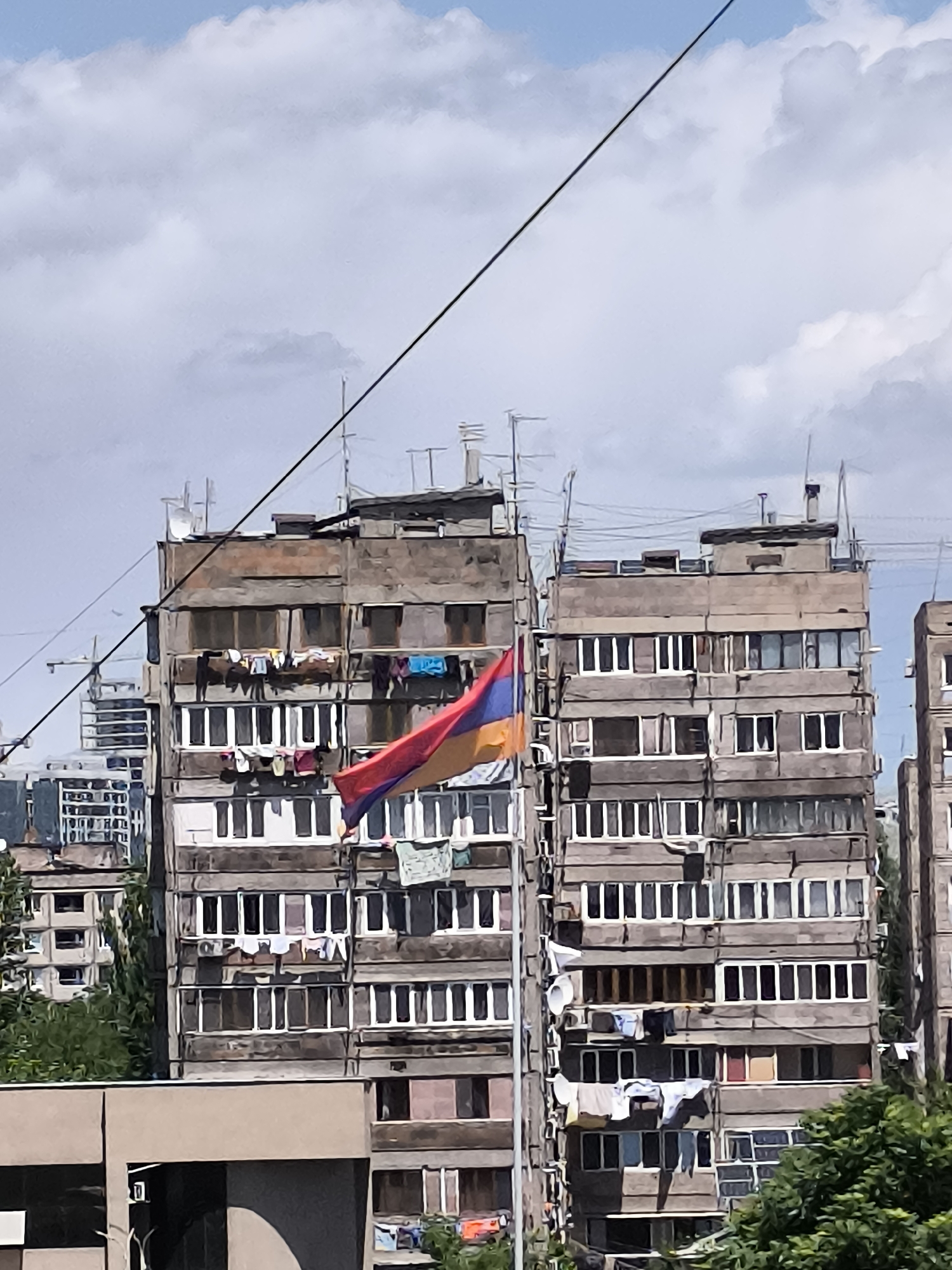 This image captures a vibrant urban scene in Yerevan, Armenia, featuring several multi-story residential buildings under a partly cloudy sky. The architecture suggests typical Soviet-era concrete apartment blocks, characterized by numerous windows and balconies.
The main subjects are these large, somewhat aged buildings. They show signs of active habitation, with many windows updated to modern white frames, and various clotheslines hanging laundry from balconies and ledges, indicating daily domestic life. On the rooftops, an array of antennas and satellite dishes are visible, suggesting television and internet access.
A prominent Armenian flag, displaying its horizontal red, blue, and orange stripes, hangs from one of the central buildings, catching the light and unfurling slightly in a breeze. This flag is a key identifier of the location.
The activity depicted is primarily the mundane yet essential aspect of urban living, with clothes drying in the open air. No people are visible, but the signs of their presence are abundant.
Notable details include the bright daytime lighting, suggesting clear visibility and good weather despite the clouds. A thick black cable or power line stretches diagonally across the upper portion of the frame, adding to the urban feel. Green foliage from trees is visible at the lower edges of the image, providing a natural contrast to the concrete structures.
There is no discernible text visible in the image. The overall impression is one of a bustling, lived-in urban environment.