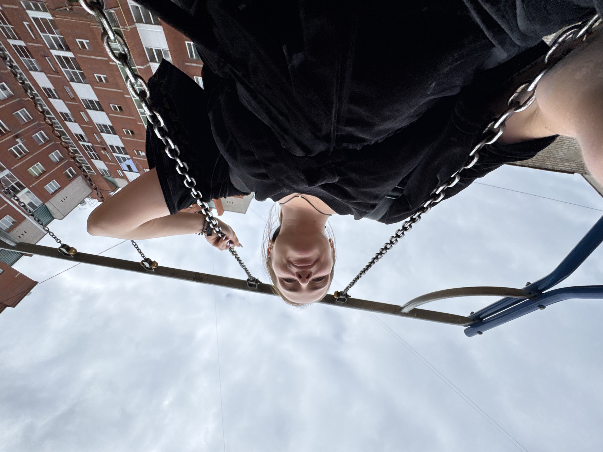 A young woman is photographed from a low angle, upside down, while sitting on a swing set. She is wearing a black velour top and has a choker necklace on. Her hands, adorned with bracelets and painted nails, grip the chains of the swing. She is smiling at the camera, her expression suggesting joy or playfulness. In the background, a tall, brick apartment building with numerous windows is visible, indicating an urban or suburban residential area. The sky above is overcast, suggesting a cloudy day. The scene appears to be outdoors, likely in a playground or park. The orientation of the photo implies the photographer is also on a swing, looking up at the woman. There is no visible text in the image. The overall mood is one of casual fun and a moment captured spontaneously.