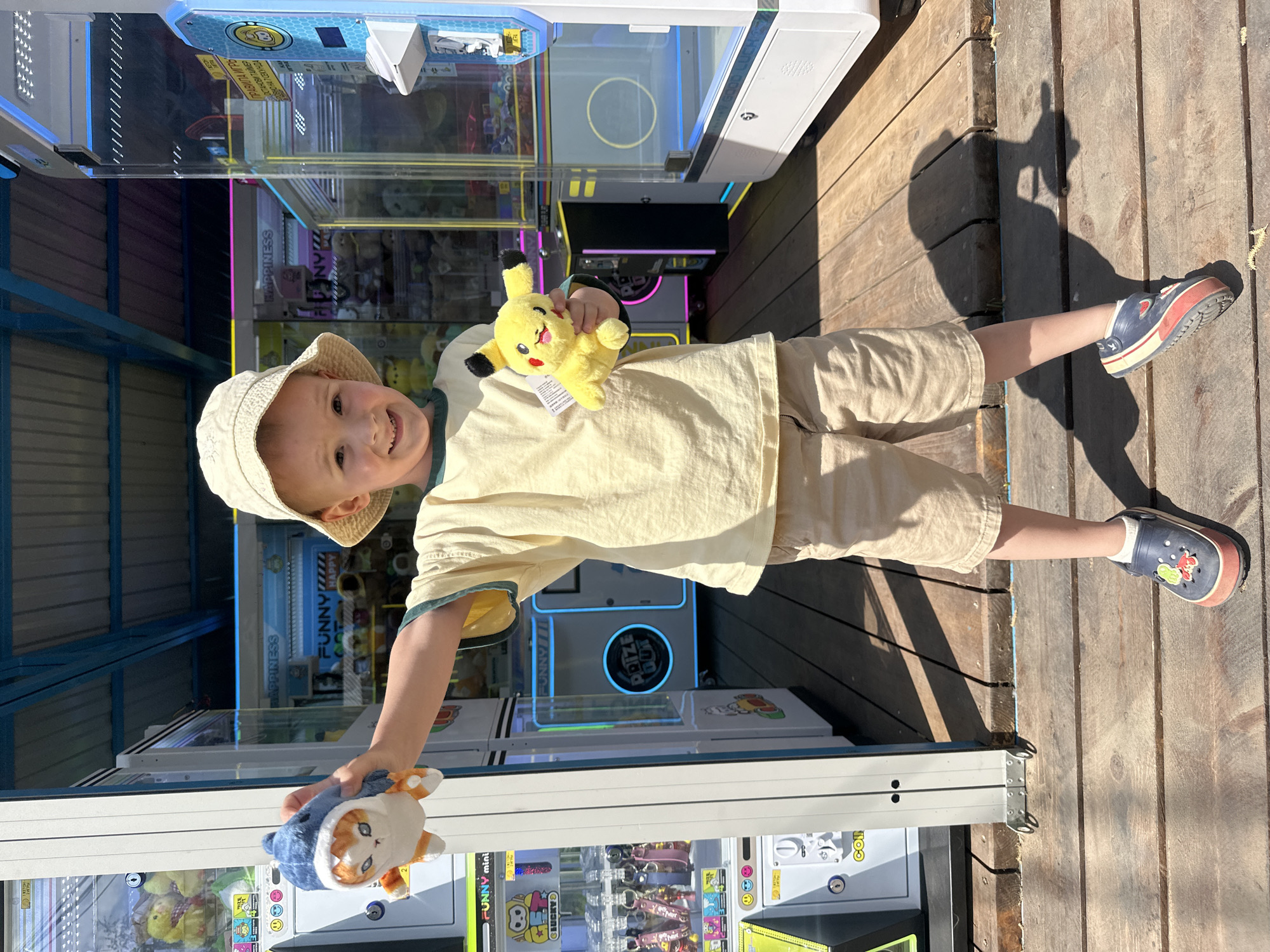 A young child stands in an arcade, holding a Pikachu plush in one hand and a plush animal in the other. The child is wearing a light-colored bucket hat, a yellow shirt with green trim, and khaki shorts. Their footwear consists of blue and red clogs. The child is surrounded by various claw machines. The ground appears to be wooden planks. The location seems to be outdoors with a partial view of the sky and roof.