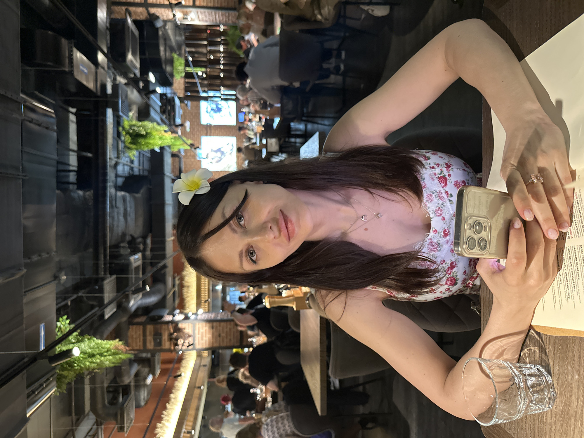 A woman with long dark hair sits at a table in a restaurant. She is wearing a floral dress and has a white flower in her hair. She is holding a smartphone in her hands. On the table are a glass of water and a menu written in Russian. In the background, other patrons are visible, along with exposed brick walls and industrial-style ductwork with greenery attached. This suggests a modern or renovated industrial interior. The OCR text indicates a menu with dishes like Carpaccio and Caesar Salad. The location is likely a restaurant in Russia given the menu in Russian and the context of the image. The presence of the word "Saint Petersburg" cannot be determined from the image.