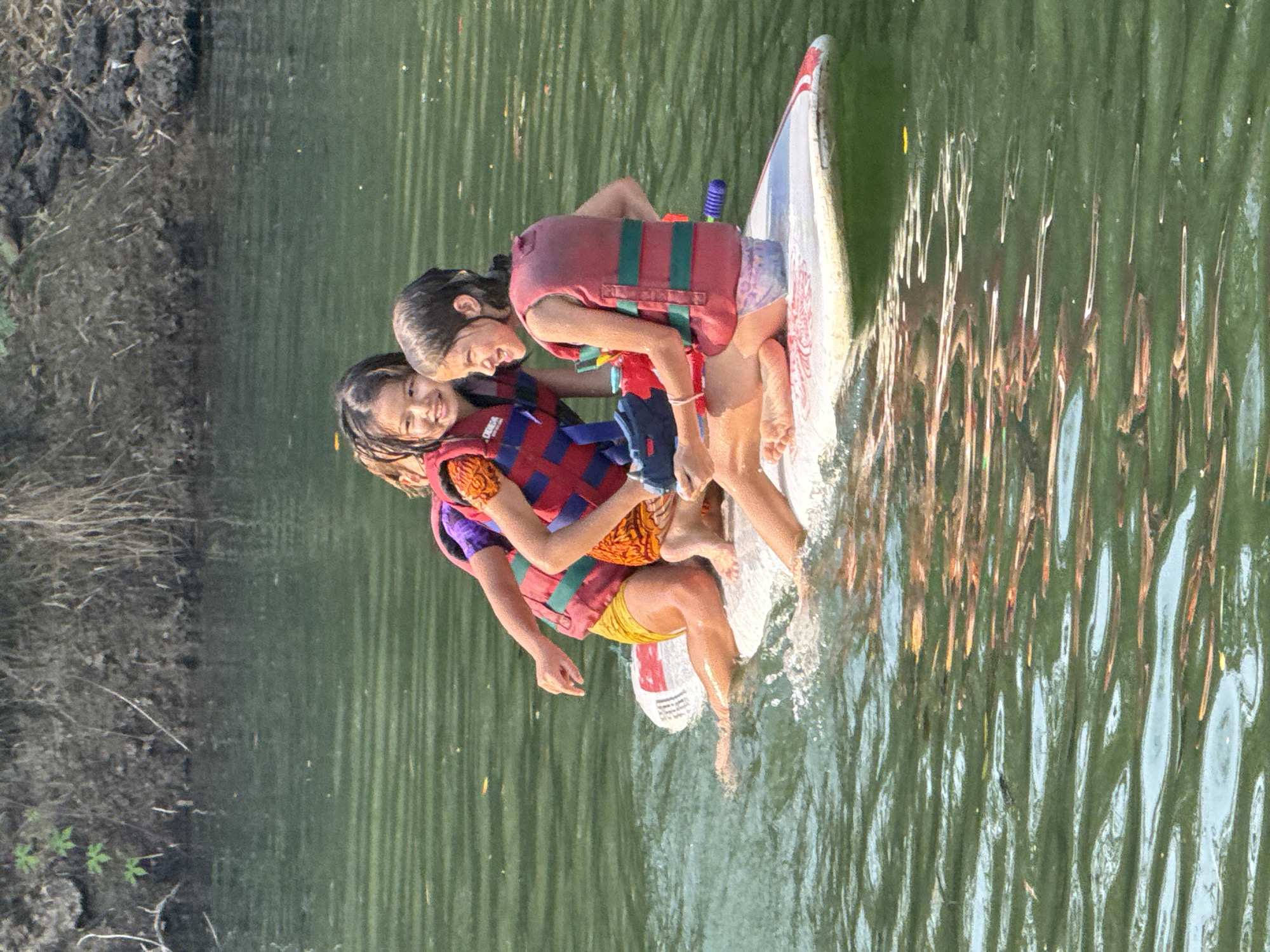 Two young girls wearing life vests stand on a white paddleboard in a body of water. The water reflects light and creates wavy patterns. The background shows vegetation along the shoreline. This appears to be Tambon Nam Phrae, Thailand.