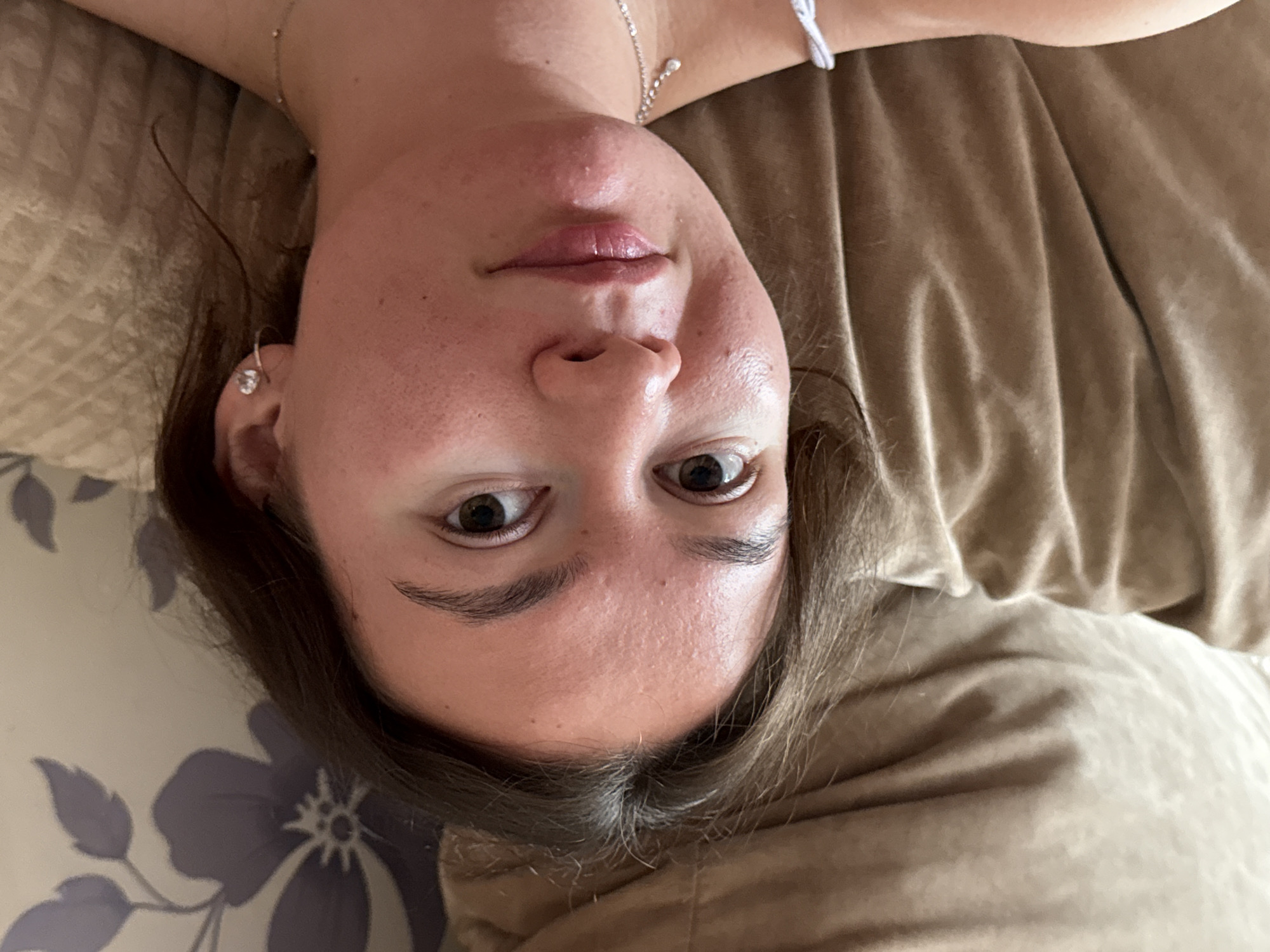 A close-up, head-and-shoulders shot from a slightly low angle shows a young woman with fair skin lying on a bed. The background consists of floral-patterned bedding to the left and a plain brown duvet or pillow to the right. The woman's dark brown hair is parted in the middle and falls around her face. She has brown eyes, a small nose, and slightly parted lips. She is wearing a silver necklace and a small earring in her left ear. No specific details indicate a location in Chaikovsky, Russia. The environment suggests a domestic setting, likely a bedroom.