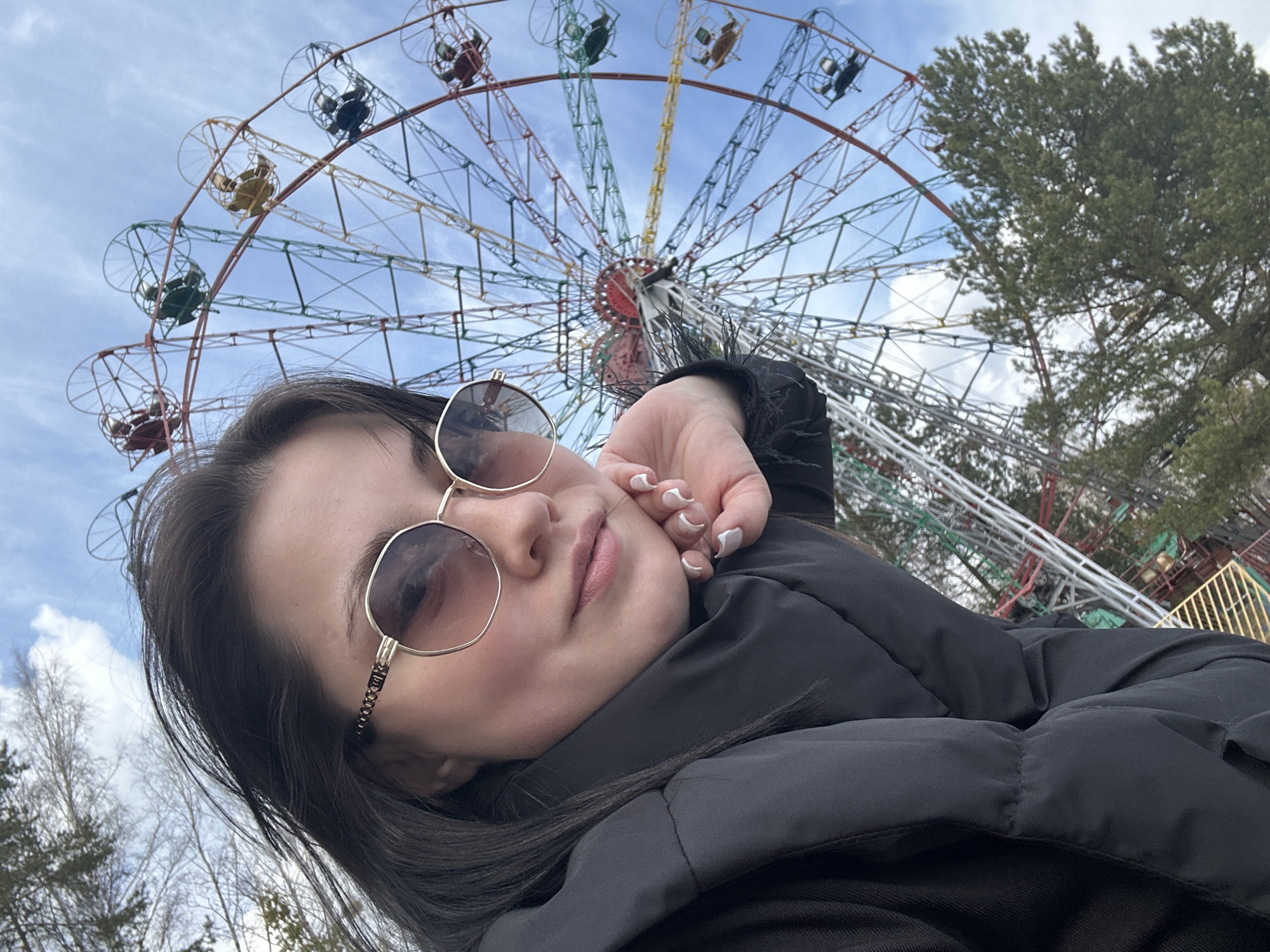 A low-angle selfie features a young woman in the foreground, partially obscuring a large Ferris wheel behind her. The woman has dark hair and is wearing a black puffer jacket and gold-framed sunglasses. Her left hand is raised near her face, revealing nails with white French tips.
The dominant feature in the background is a large Ferris wheel, its complex metal framework painted in various colors. Multiple cabins, distinguishable by red, green, yellow, and blue hues, are visible along its circumference, some appearing to have occupants. The sky above is bright and partly cloudy, suggesting it is daytime. A large evergreen tree is present on the right side of the image, while some bare deciduous trees appear on the left, indicating a season like late winter or early spring.
The setting is an amusement park or fairground in Chaikovsky, Russia. The woman's expression is neutral, and the overall atmosphere seems relaxed. There is no visible text in the image.
