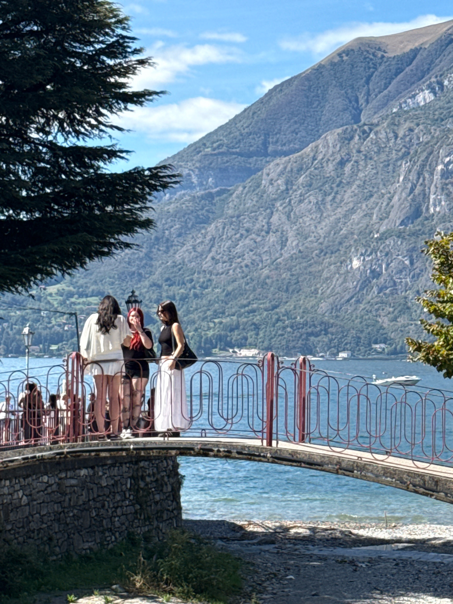 The image depicts a scene in Gittana, Italy, featuring a stone bridge spanning a body of water. On the bridge, three young women stand facing away from the camera, engaged in conversation. The woman on the left wears a white outfit, the central woman has red hair and is wearing a black top and shorts with patterned stockings, and the woman on the right is in a black top and a long white skirt. The bridge's railing is made of red metal with an oval pattern. Behind the women, the expansive blue water of a lake stretches out, with a white boat visible in the distance. The backdrop consists of steep, forested mountains under a bright blue sky with scattered white clouds. A large tree with dark green foliage partially obscures the upper left portion of the image. The bridge's stone foundation is visible on the lower left, and the foreground shows a rocky, pebbly shore with some sparse green vegetation.