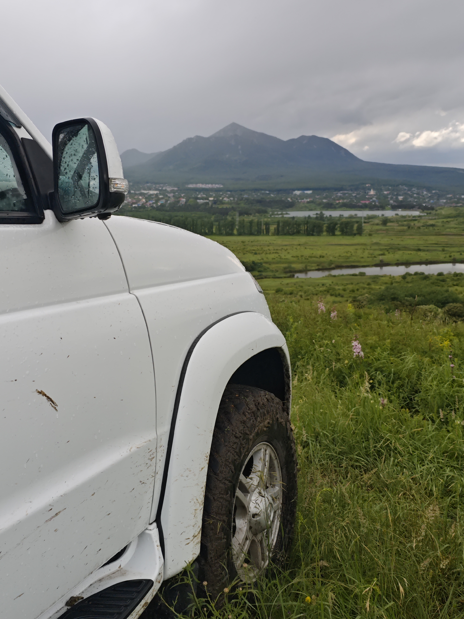 The image shows the front of a white vehicle, possibly an SUV, with a dirty tire indicating off-road use. The side mirror has water droplets, suggesting recent or ongoing rain. The background reveals a landscape including a green field with tall grass, a river or waterway, and a town or settlement in the mid-ground. A mountain is visible in the distance under a cloudy sky. This is Inozemtsevo, Russia.