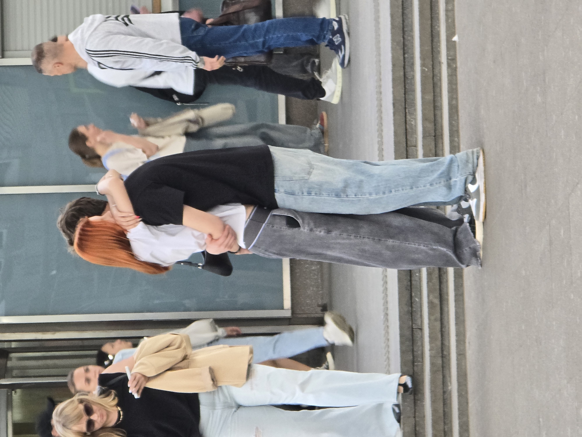 A medium outdoor shot captures a group of people on a sidewalk. An embracing couple stands out in the foreground, while other pedestrians are behind them. The backdrop includes a building facade with large windows, and the edge of a staircase. The location is identified as Khimki, Russia.