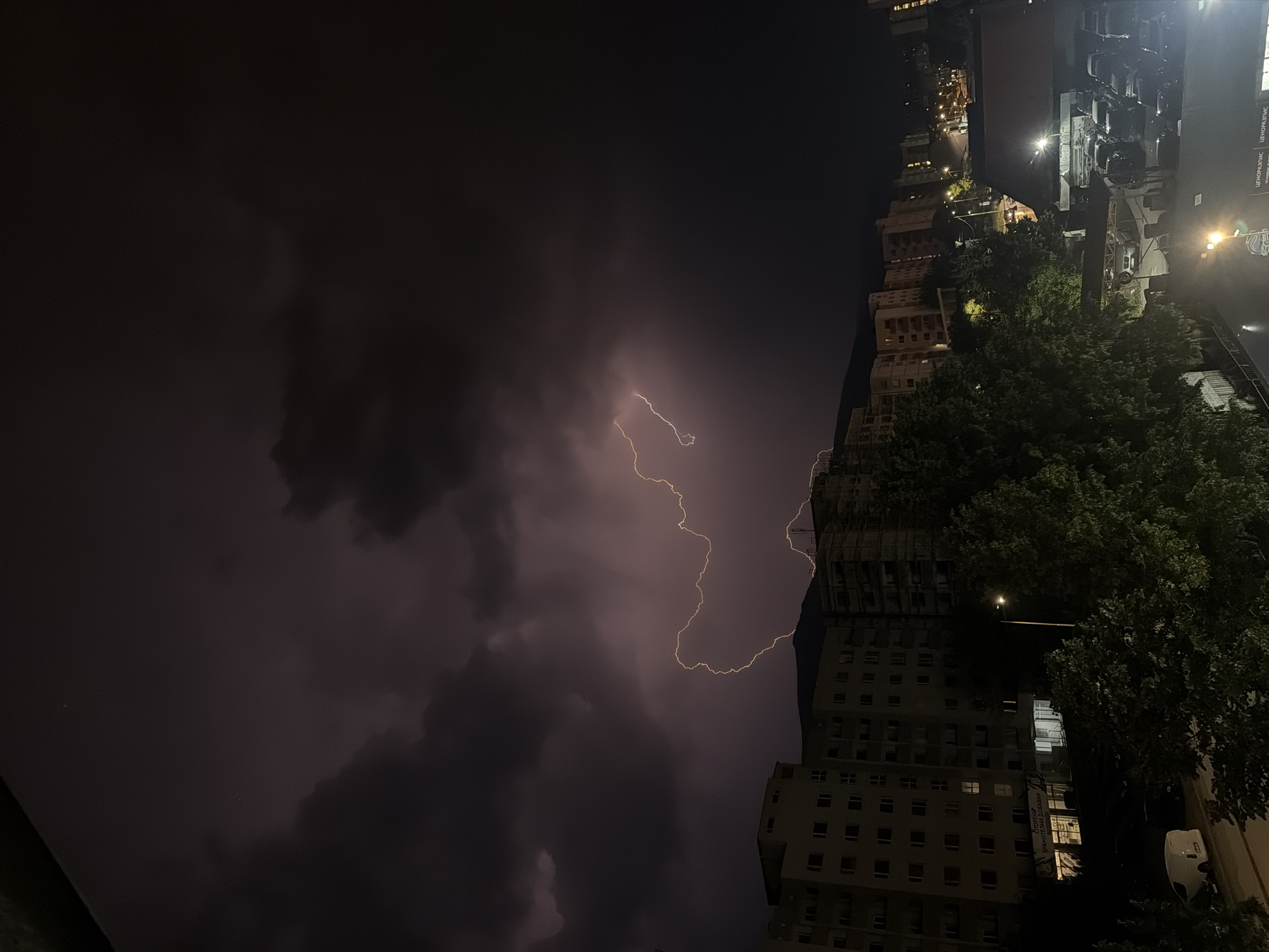 This image captures a dramatic thunderstorm over Sofia, Bulgaria, at night. A jagged bolt of lightning illuminates the dark, ominous clouds, casting a faint purple glow. Below the storm, the cityscape is visible, with apartment buildings and trees silhouetted against the night sky. The lightning appears to be striking behind a mountain range in the distance. In the foreground, the lights of the city can be seen, including streetlights and illuminated windows in the buildings. There are no people or animals clearly visible in the image. The scene depicts a powerful natural event occurring over an urban environment.