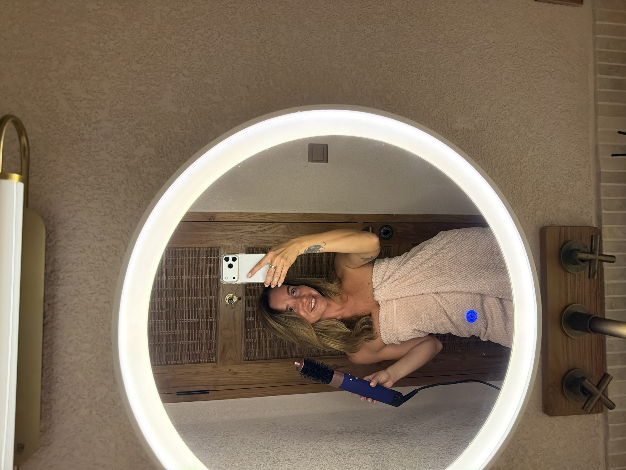 A woman is taking a mirror selfie in a bathroom, holding a blue hairdryer and wrapped in a pink towel. She is standing in front of a wooden door and a textured wall. A large, circular mirror with a bright light ring is centered in the image. The scene appears to be in a hotel bathroom in Mengwi, Indonesia.