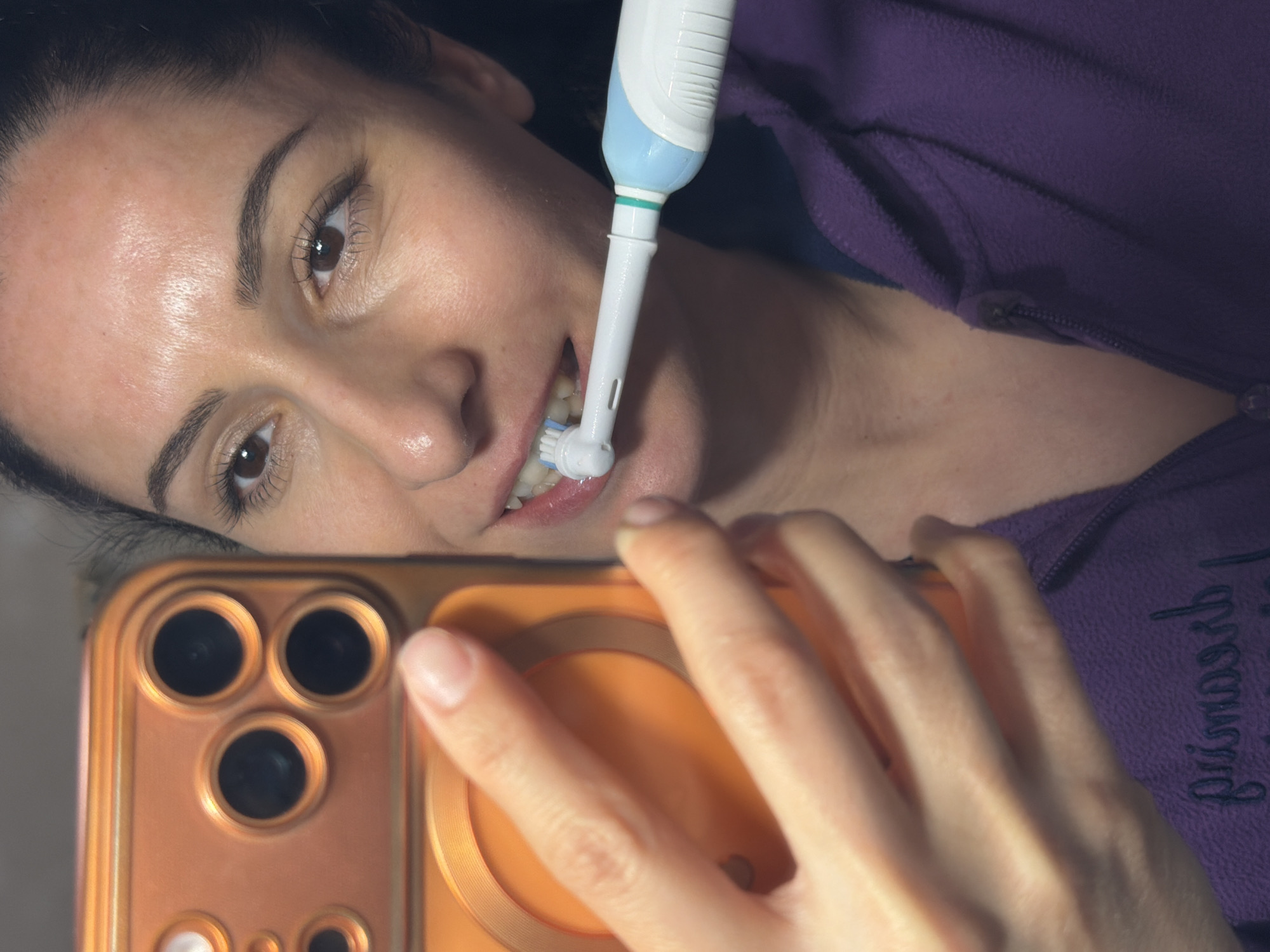 A woman is taking a selfie while brushing her teeth with an electric toothbrush. She is holding an orange-colored smartphone, possibly an iPhone, with its camera facing her. She is wearing a purple zippered fleece top. The location is not discernible in the provided image and therefore, cannot be placed in the city of Pleven, Bulgaria.