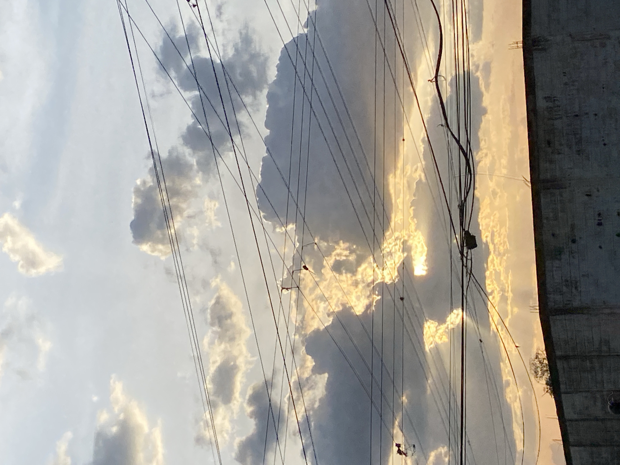 The image shows a section of the sky in Tinaquillo, Venezuela, with scattered clouds obscuring the sun, giving the sky a golden hue. Several power lines crisscross the view. A concrete structure is visible on the right side of the frame.