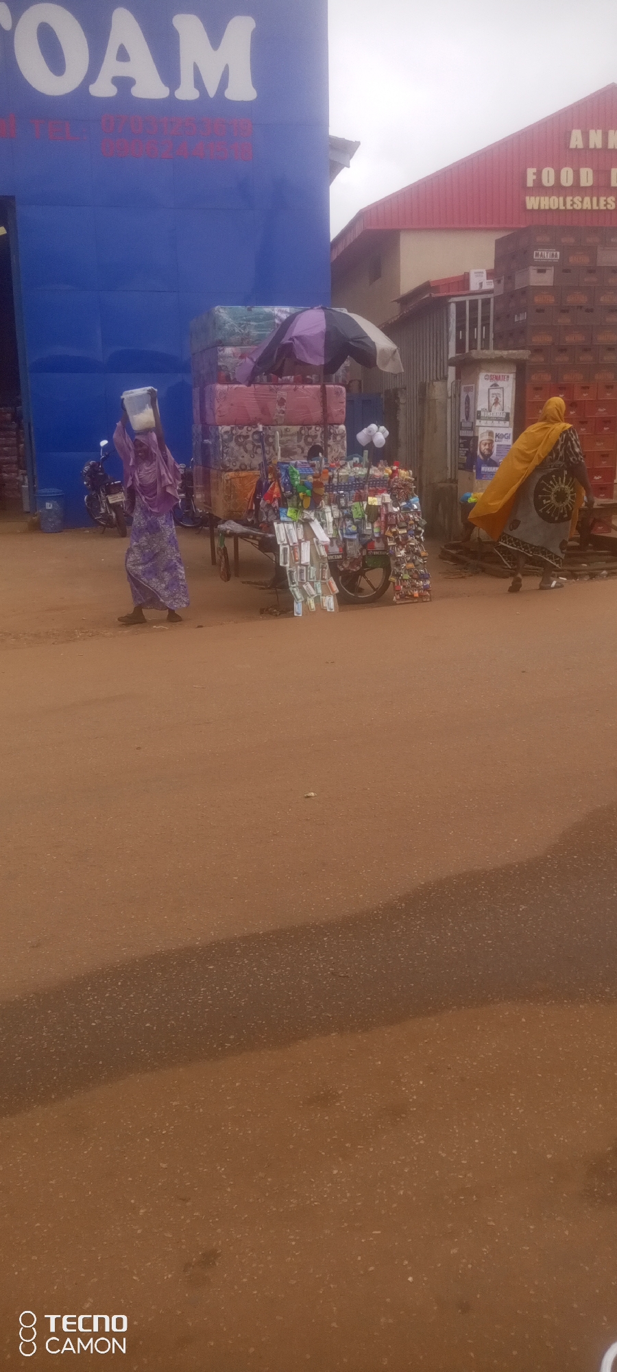 In Ankpa, Nigeria, a street scene unfolds with a blue building on the left displaying the word "FOAM" in large white letters, followed by contact telephone numbers. In front of the building, a stack of mattresses is visible. To the right, a woman in a purple abaya and headscarf carries a white bucket on her head, walking towards the left. Adjacent to the mattresses, a street vendor's cart is laden with various small items, including packaged goods and what appear to be keychains or small accessories, under a striped umbrella. Further to the right, a red building with "ANKP..." and "FOOD D..." signage is partially visible. A pile of boxed goods, possibly beverages, is stacked against this building. Another person, wearing a yellow headscarf and a patterned dress, walks away from the camera toward the right. The ground is a dusty, reddish-brown color, indicating a dry, outdoor environment. The sky is overcast.