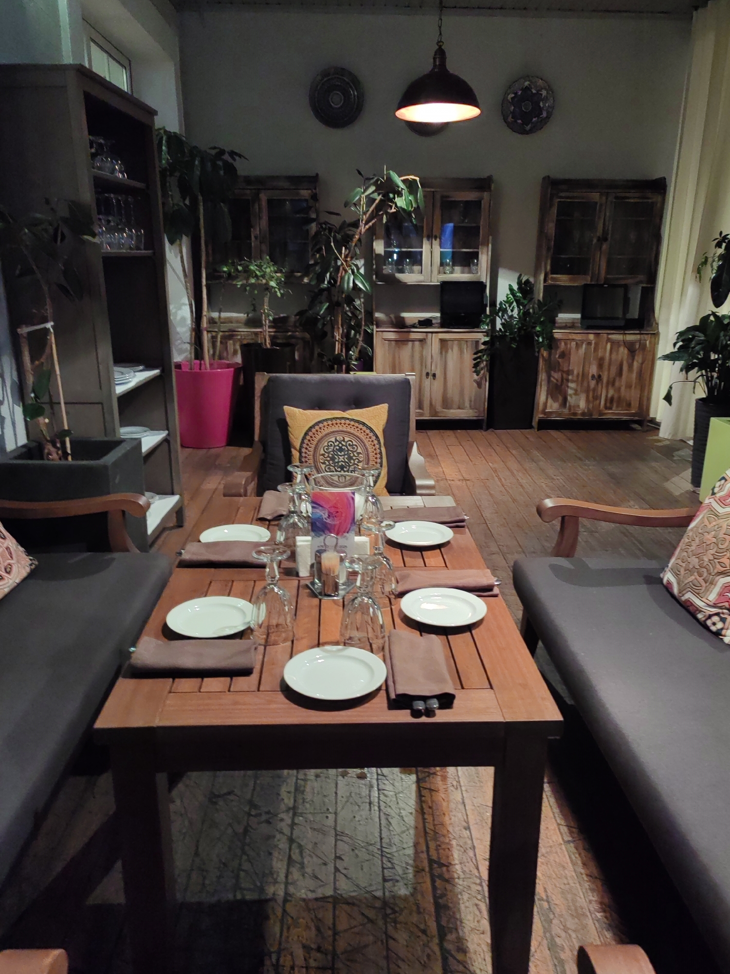 The image depicts an indoor dining area, likely a restaurant. A long, wooden table is set with plates, cutlery, and glassware. Two cushioned benches or seating areas flank the table. In the background, there are wooden cabinets and shelves filled with decorative items and possibly cookware. Several potted plants are scattered throughout the room, adding a touch of greenery. The lighting is dim, with a hanging lamp providing illumination. The overall aesthetic suggests a cozy and rustic ambiance. The location is not provided in the image.