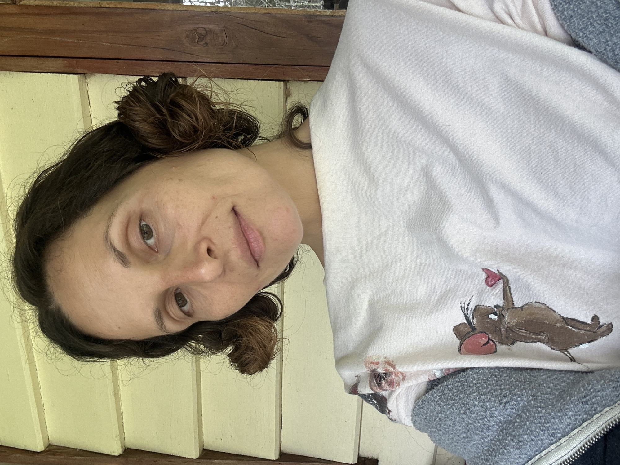 A woman with dark brown hair, styled in two buns, faces the camera. She wears a light-colored t-shirt with a cartoon mouse graphic holding a heart on the left side of the chest, and a grey zippered jacket. The background consists of light yellow wooden planks and a dark wooden post. The image appears to be taken outdoors. There is no information available to determine the location in Thora, Australia.