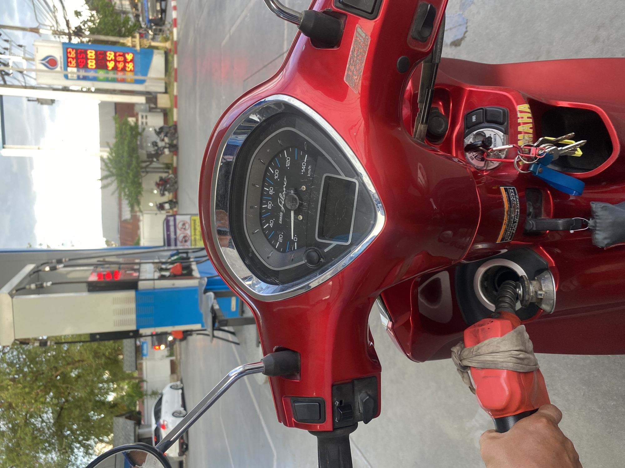 The image depicts the viewpoint from the rider of a red Yamaha Grand Filano scooter at a gas station in Baan Mae Nam, Thailand. The scooter's dashboard shows the speedometer with a needle pointing to a low speed. Keys dangle in the ignition. The rider is holding a red gas pump nozzle with a rag tied around it, filling the scooter's gas tank. In the background, a gas price sign displays prices in Thai Baht. Gas pumps and the station's structure are visible, along with a few other vehicles parked in the distance.