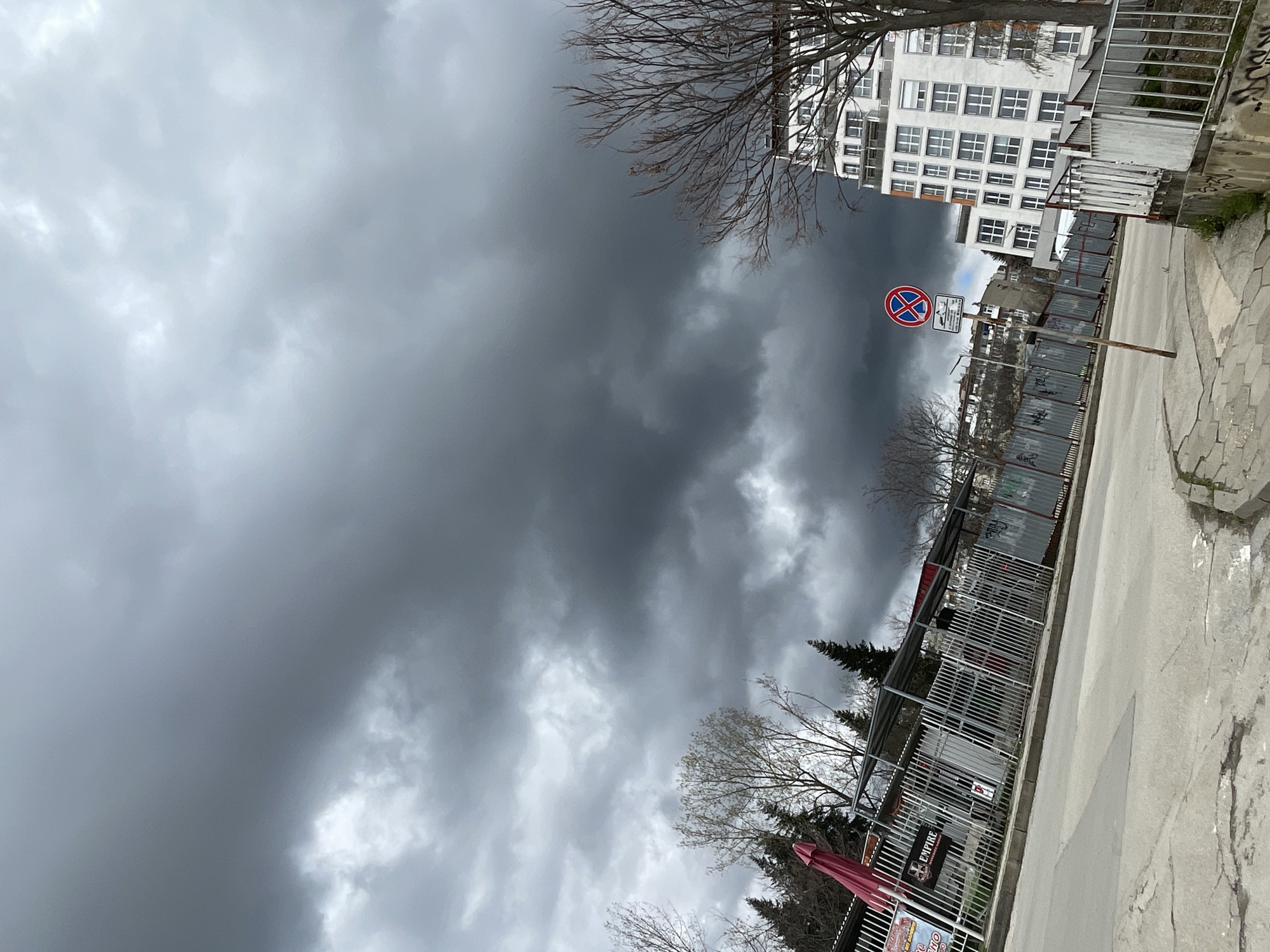 The image shows a street scene in Veliko Tarnovo, Bulgaria on an overcast day with a sky full of dark clouds. A building with multiple windows is visible on the right, along with a street featuring a sidewalk. A “No Stopping” sign stands near the building. A metal fence runs along one side of the street, near an outdoor business, partially obscuring some trees.