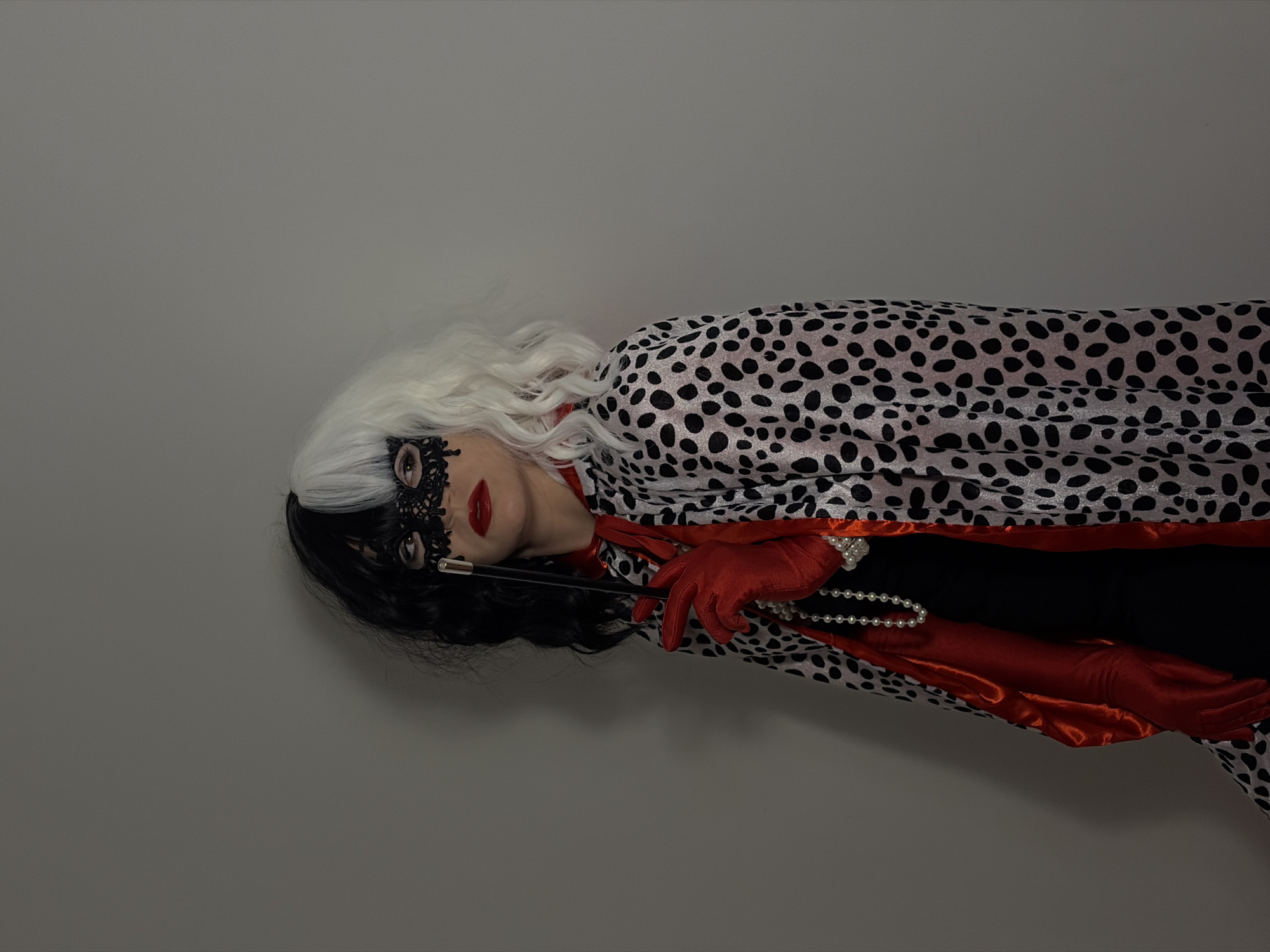 A person dressed as Cruella De Vil is seen indoors. The person has half black and half white hair, wears a black lace mask, and is wearing red lipstick. The person is also wearing red satin gloves, a pearl necklace, and a dalmatian-print cloak with a red satin lining. The person is holding a cigarette holder. The background is a plain grey wall. There is no evidence of the location being in Budva, Montenegro.