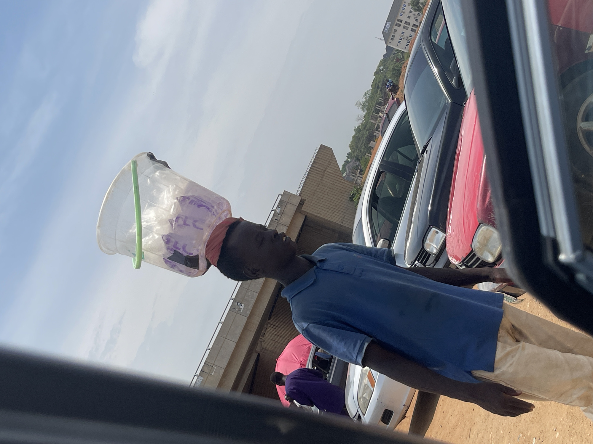 This image captures a street scene in Galadimawa Village, Nigeria, from the perspective of a vehicle. The photograph is notably tilted, giving a candid and dynamic feel.
The central subject is a young person, likely a boy or young man, depicted from the waist up. He is wearing a blue short-sleeved polo shirt and light tan or khaki pants. He skillfully balances a large, clear plastic container, possibly a water dispenser or a bucket containing goods, on his head. The container is secured by a reddish-pink cloth band around his forehead. His expression is neutral as he looks forward, possibly scanning for customers or navigating the road.
In the background, several cars are visible, including a prominent red sedan, a dark-colored vehicle, and a white/silver car, suggesting a traffic-heavy road or a stopped vehicle situation. Further back, another person wearing a purple top stands near a pink umbrella, and other figures are partially visible. A concrete overpass or bridge structure spans the mid-ground, while distant multi-story buildings and greenery adorn a hill under a bright, partly cloudy sky. The lighting indicates it is daytime. The foreground shows part of a car's dark interior, such as a door frame or window, from which the photo was taken.
Some indistinct purple text, possibly a brand name like "PLC," can be seen on the clear plastic container on the subject's head, but it is not clearly readable. The image portrays a common sight of street vending in many Nigerian urban and semi-urban areas.