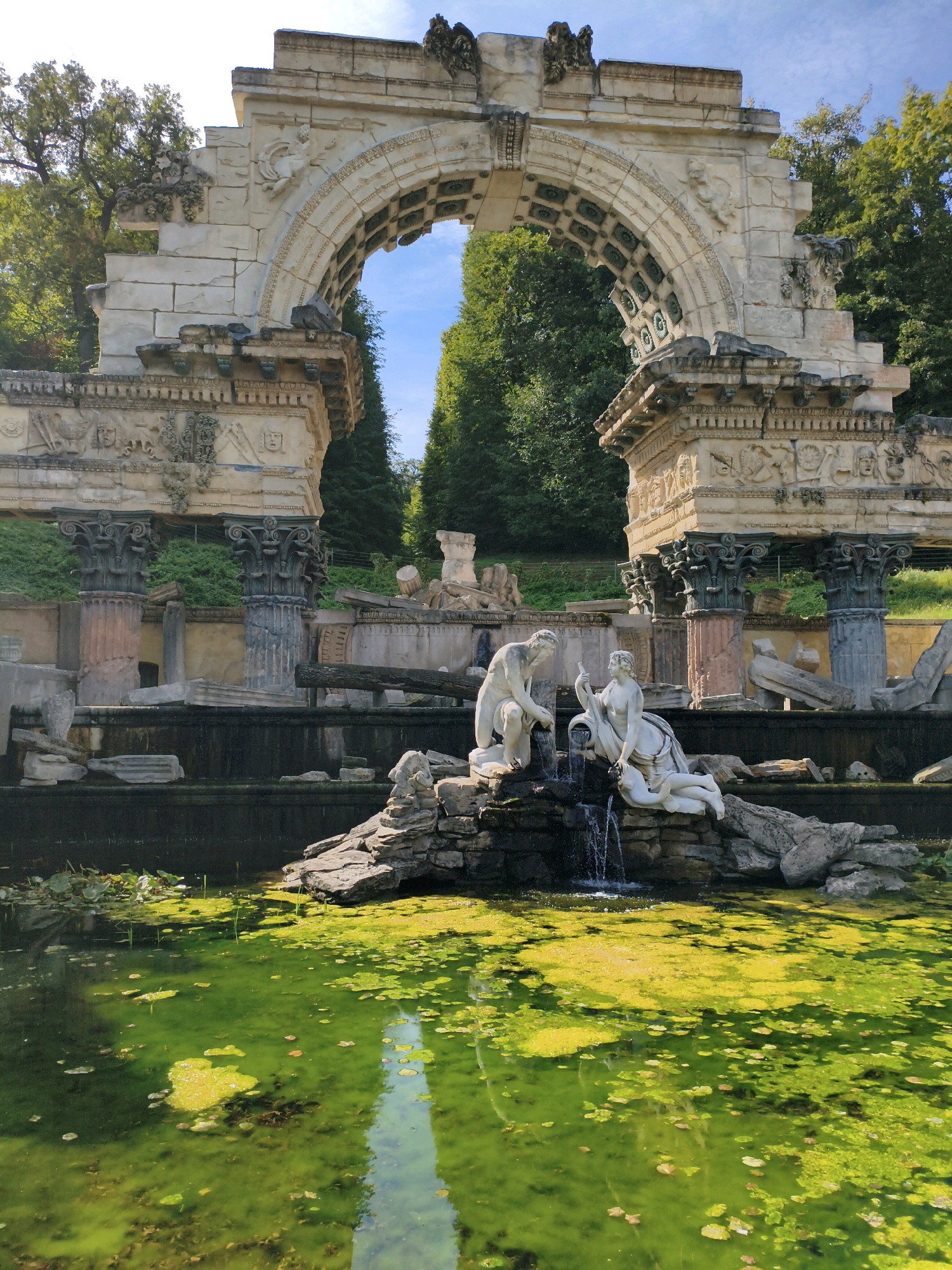 The image depicts the Roman Ruin in Schönbrunn Palace Gardens, Vienna, Austria. It features a large, weathered stone archway with intricate carvings, suggesting ancient Roman architecture. In the foreground, a pond filled with green algae and lily pads reflects the sky. A water feature with two white marble statues is present, one male and one female, seemingly interacting with the water. The statues are positioned on a rocky outcrop where water cascades down. The background is composed of lush green trees and a clear blue sky. The overall scene conveys a sense of historical grandeur and natural beauty.