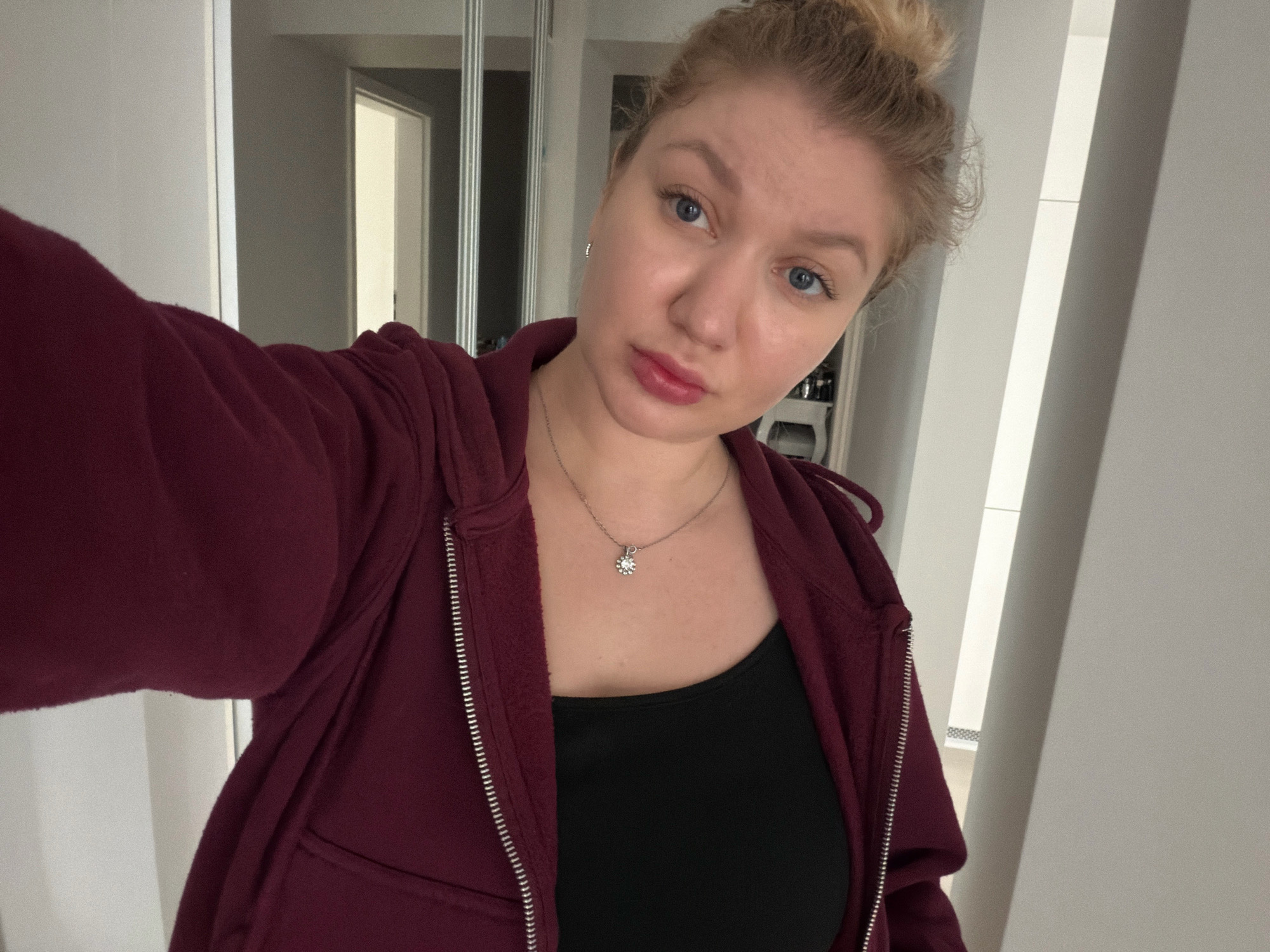 A young woman with blonde hair pulled into a bun, blue eyes, and light skin is taking a selfie. She is wearing a dark red zippered hoodie over a black tank top and a silver necklace with a small pendant. She has light makeup on, including lip gloss. She is standing indoors in what appears to be a residential hallway with white walls and subtle gray accents. In the background, there is a glimpse of an open doorway leading to another room and a tall, narrow mirror or closet with vertical silver stripes. The setting suggests an interior residential space in the city of Koszalin, Poland, possibly a hallway in an apartment building or a house.