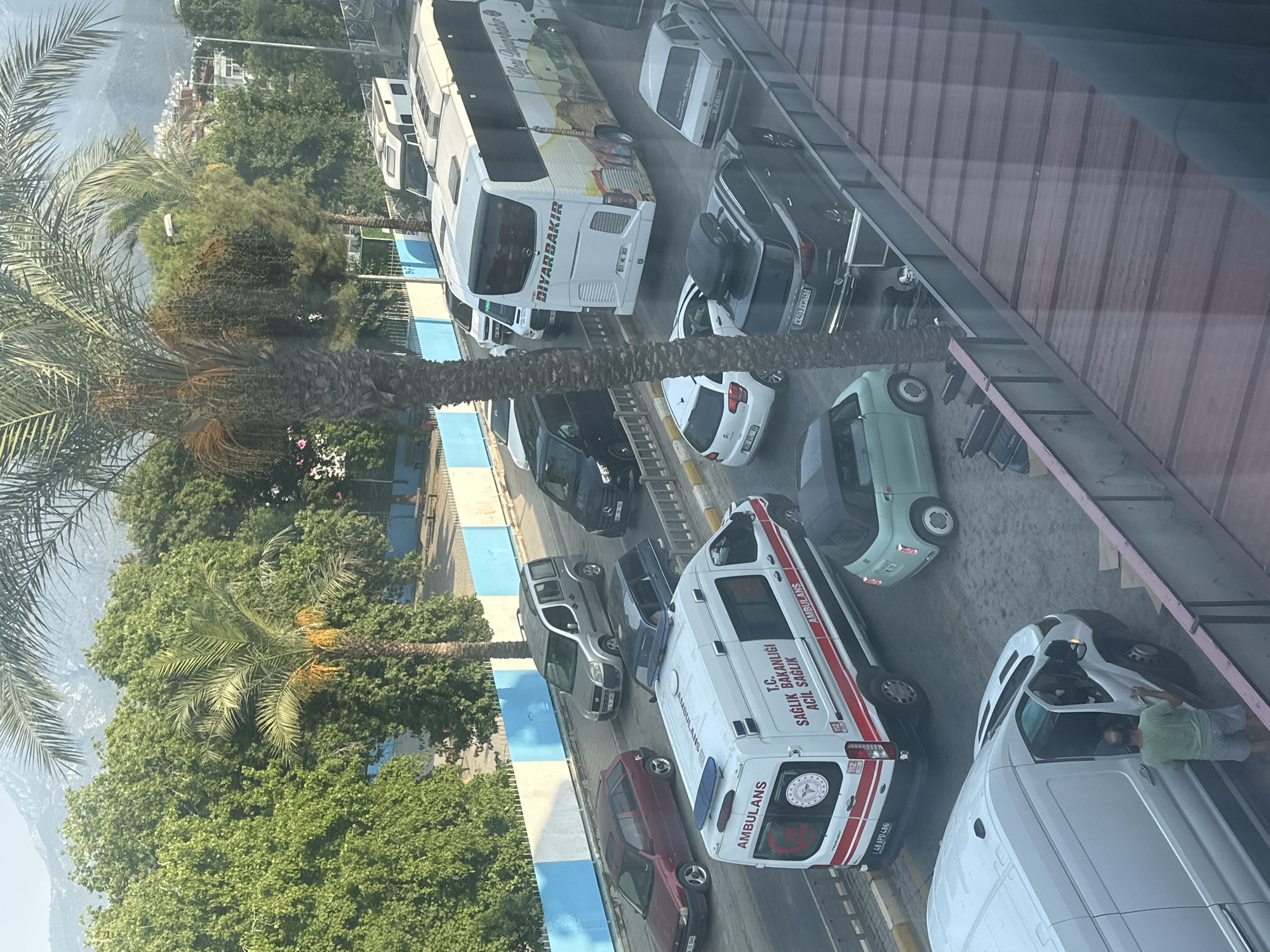 The image depicts a street scene in Fethiye, Turkey, with a variety of vehicles. In the foreground, a white van has its driver's side door open, and a man in a green t-shirt and grey shorts is standing next to it, appearing to be interacting with the vehicle. Behind the van, an ambulance with "AMBULANS" and "T.C. SAĞLIK BAKANLIĞI ACİL SAĞLIK" (Republic of Turkey Ministry of Health Emergency Health) written on its side is stopped in traffic. To the right of the ambulance is a small, light green car.
Further down the street, a large white bus with "DIYARBAKIR" written on its back is prominent, alongside a dark-colored SUV with a rooftop cargo carrier. Several other cars, including a red hatchback, a silver multi-purpose vehicle, and a black car, are also visible in the traffic jam. The environment includes palm trees, lush green foliage, and some buildings in the background, suggesting an urban or semi-urban setting. The sky is bright, indicating daytime.