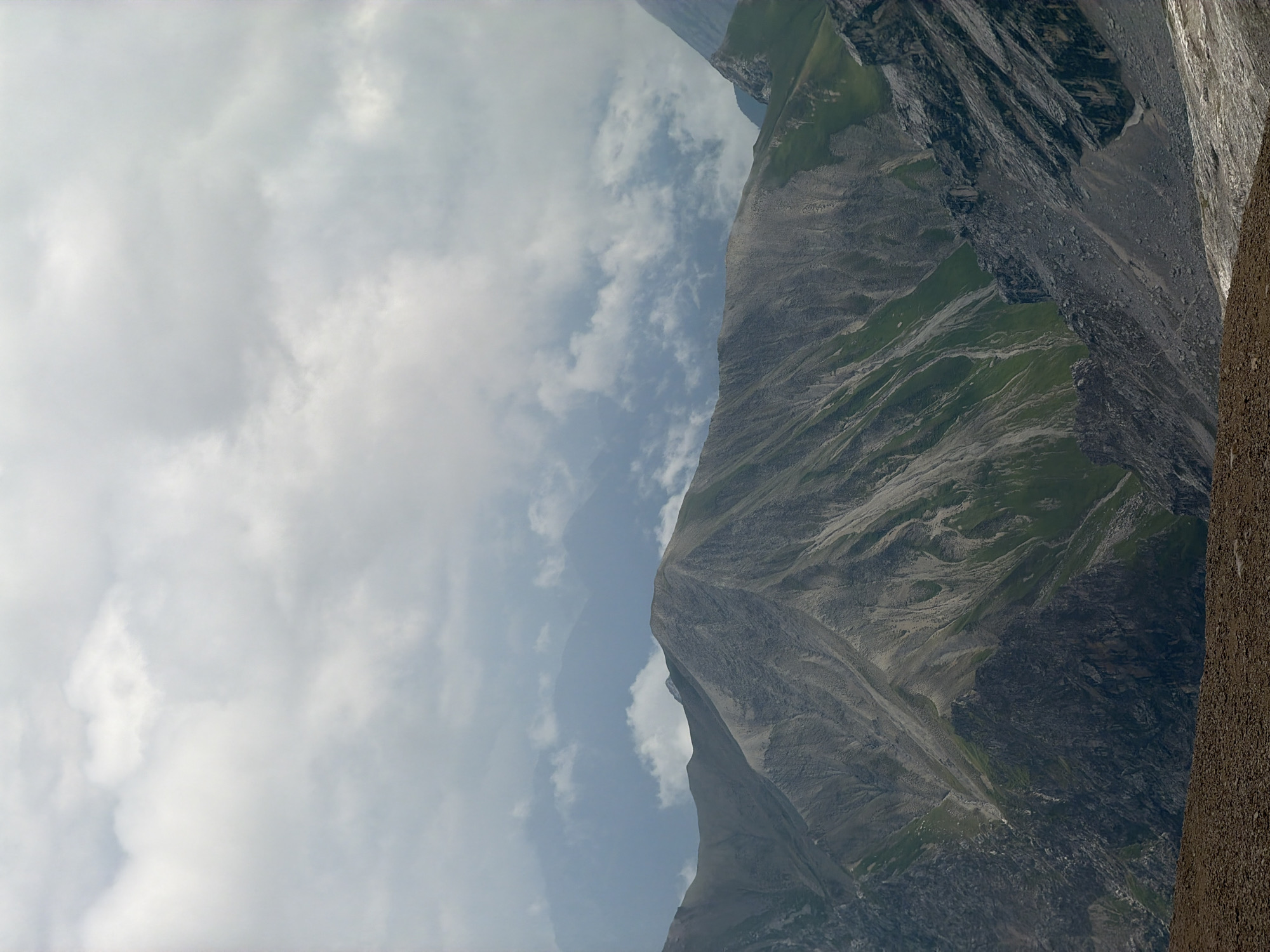 The image displays a mountainous landscape with rugged terrain and a cloudy sky. In the foreground, there is a patch of brown, rocky ground. Behind this, a large mountain slope dominates the view, characterized by steep inclines, exposed rock, and patches of green vegetation. The mountain range extends into the background, with several layers of peaks fading into a hazy, blueish distance. The sky is filled with heavy, gray clouds, obscuring any direct sunlight and creating an overcast atmosphere. The scene evokes a sense of natural grandeur and remoteness, typical of alpine environments. While the image depicts a natural setting, it does not contain any identifiable elements that can be specifically linked to the city of Garmisch-Partenkirchen, Germany. The composition focuses solely on the geological features and atmospheric conditions of the mountains.