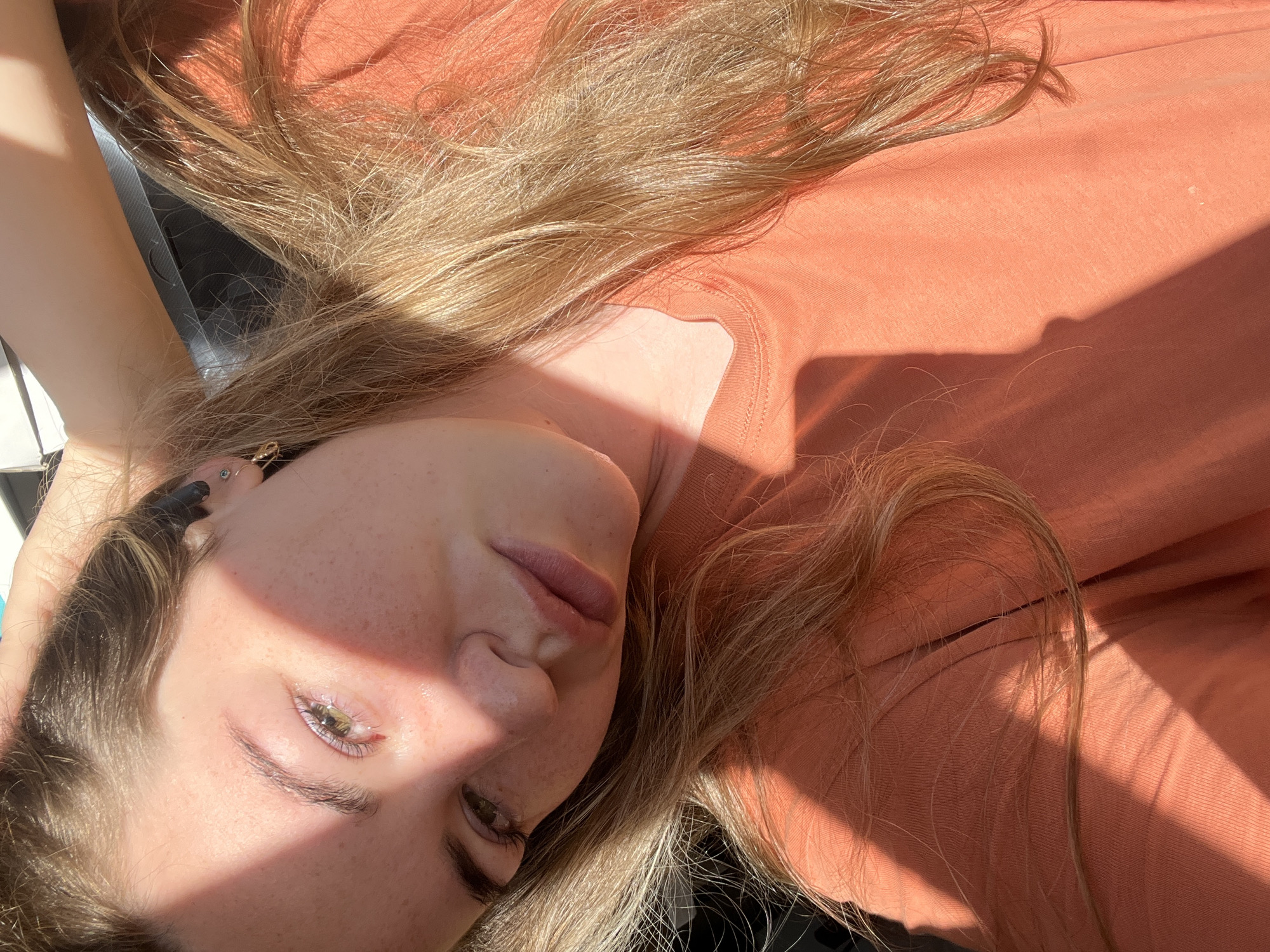 The media file is a close-up, angled shot of a young woman's face and upper body, lying down. The lighting suggests it is daytime, with strong sunlight casting distinct shadows across her skin and clothing. The woman has fair skin with freckles, light brown hair that is long and textured, and light-colored eyes. She is wearing an orange, long-sleeved shirt. A shadow, possibly from a window or blinds, creates a geometric pattern across her face and the fabric of her shirt. One ear has jewelry. The scene appears to be indoors, given the way the light is falling, but there are no clear environmental cues about the specific location beyond the assumed interior setting. The woman's expression is relaxed, with her eyes partially closed and her lips slightly parted. The overall mood is serene and casual.