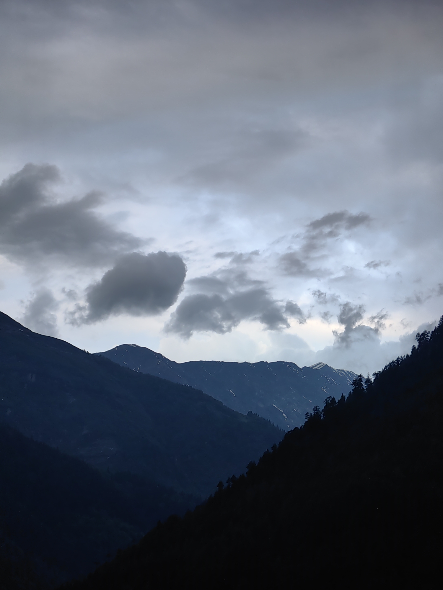 The image depicts a dramatic mountain landscape under a cloudy sky. In the foreground, a steep, dark mountainside covered in pine trees slopes downwards from right to left. Behind this, a series of mountain ranges recede into the distance, with some peaks capped with snow. The sky is filled with heavy, gray clouds, with hints of lighter sky visible in patches, suggesting either dawn or dusk. The overall mood is somber and majestic. This scene is consistent with the natural beauty found in the region of Vashisht, India, a popular hill station known for its mountainous terrain and scenic vistas.
