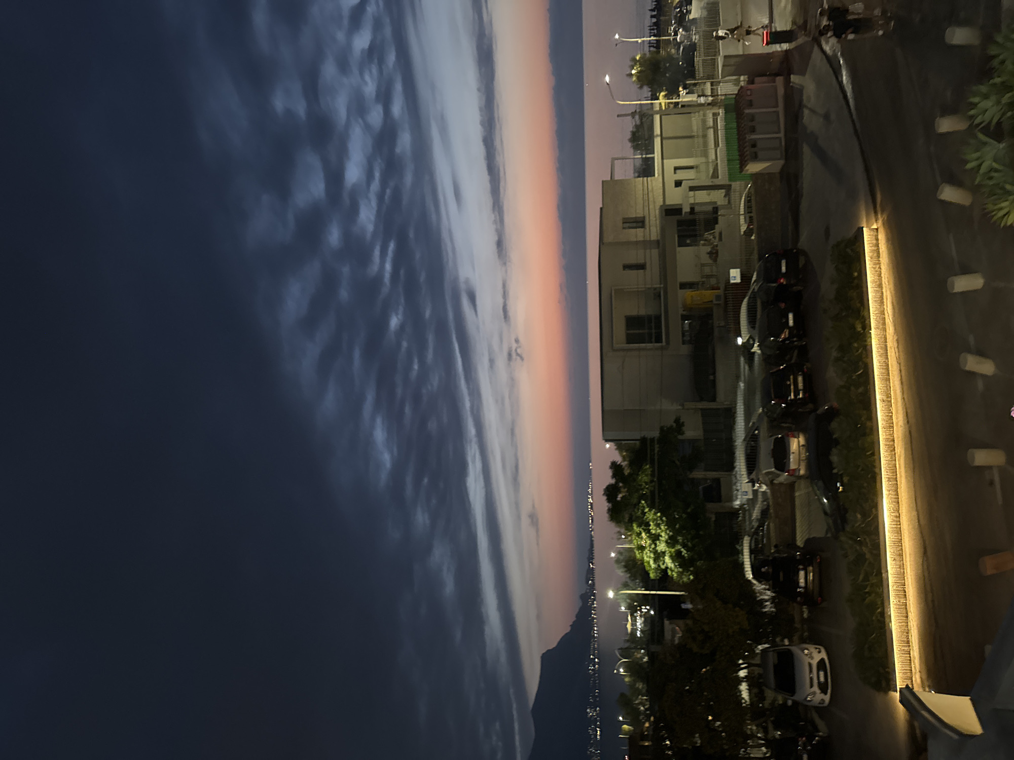 The image depicts a twilight scene in Capaci, Italy. The sky is a gradient of deep blue and dark grey clouds transitioning to lighter hues of pink and orange near the horizon, suggesting a sunset or pre-dawn glow. The sea is visible in the mid-ground, with a distant shoreline dotted with lights. In the foreground, a parking lot contains several parked vehicles, including a white hatchback on the left and several dark-colored cars. A modern building with multiple windows and balconies is situated behind the parking lot. Trees with lush green foliage are scattered throughout the scene. A paved walkway or road with embedded lighting runs along the bottom edge of the image, and several cylindrical bollards are visible on the right side of the walkway. Two figures are seen walking on the right side of the image near a trash receptacle. The overall atmosphere is calm and serene, characteristic of the coastal landscape of Capaci.