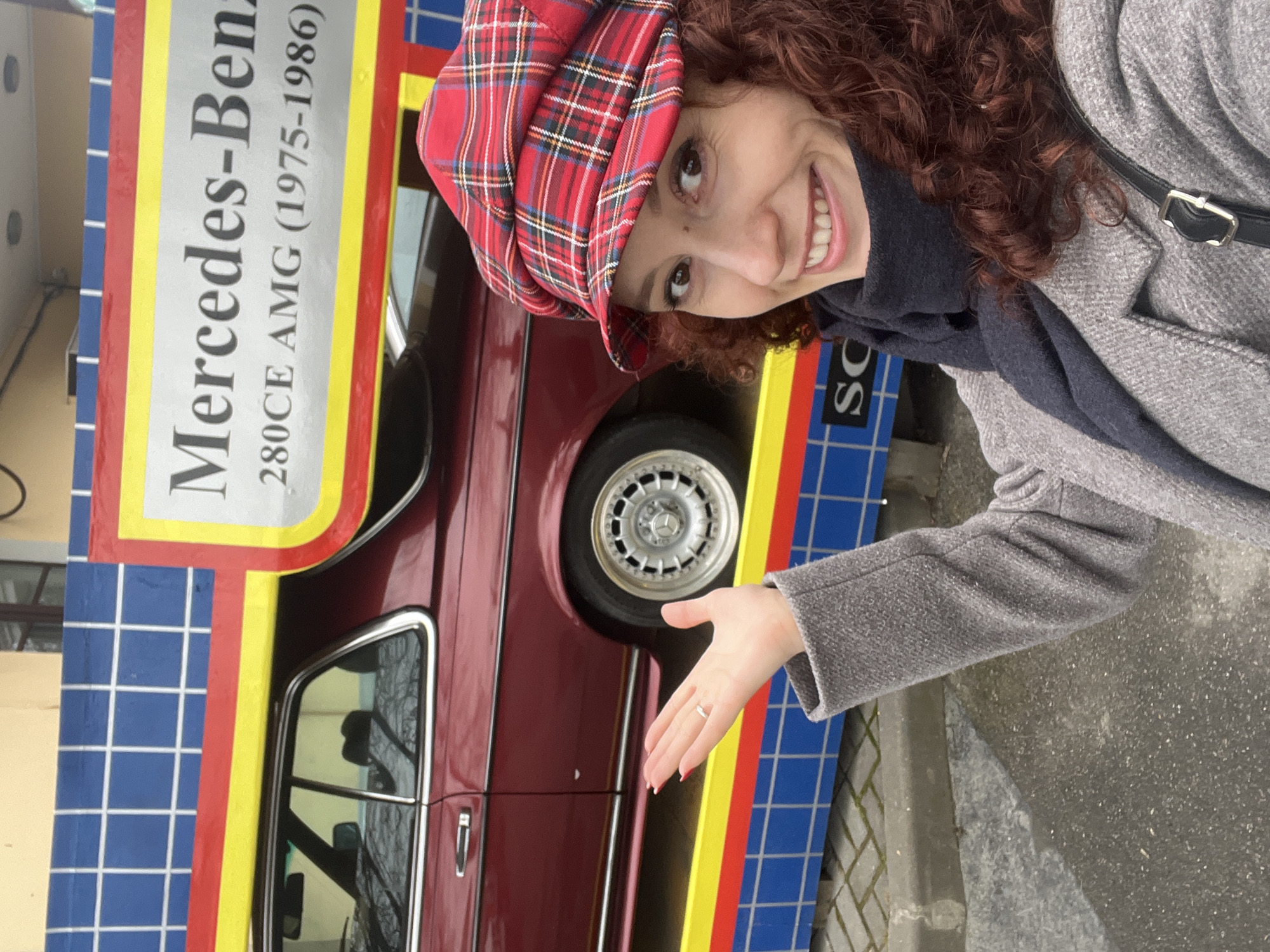 A woman with curly brown hair and a red plaid hat is posing next to a vintage Mercedes-Benz 280CE AMG (1975-1986) car. The car is a deep red color, and it is parked in front of a sign with the car's name and years of production. The sign is decorated with blue, yellow, and red stripes. The woman is wearing a gray coat and a dark scarf. She is smiling and pointing her hand towards the car. The location is most likely Saint Petersburg, Russia, as it's consistent with the architecture and street style.