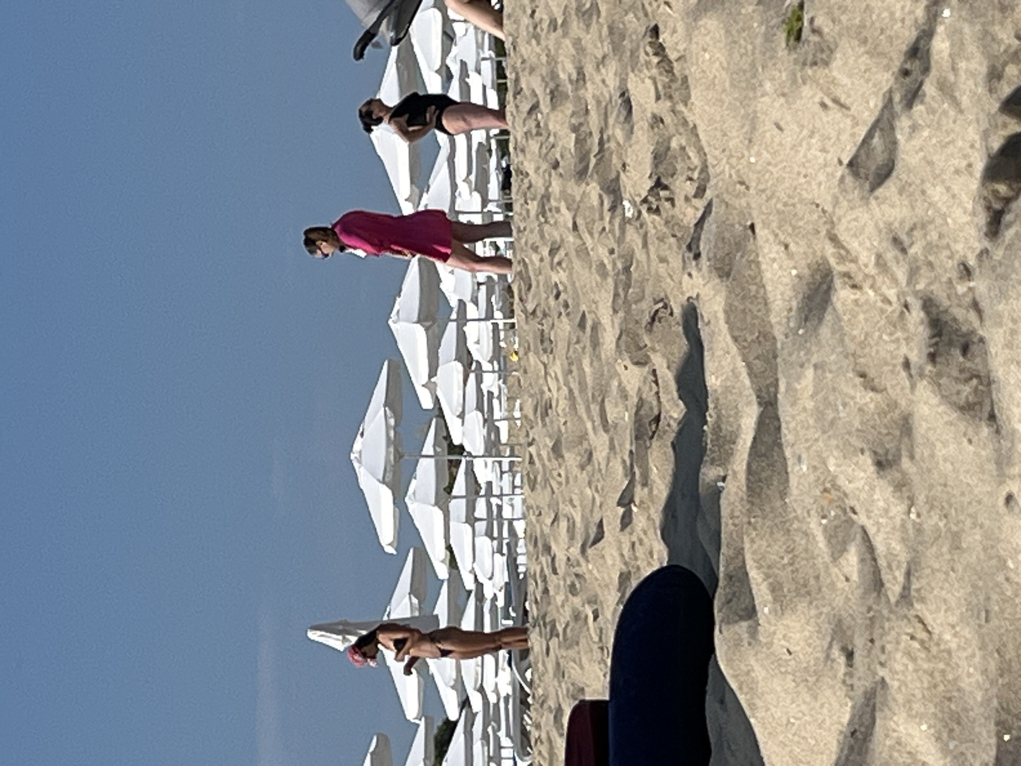 The image depicts a beach scene in Nesebar, Bulgaria, characterized by a wide expanse of sand with visible footprints and shadows. In the foreground, a dark blue cushion-like object rests on the sand. In the mid-ground, several rows of white beach umbrellas are set up, with some lounge chairs visible beneath them. Three women are present. One woman, wearing a pink hat and a dark bikini, stands facing left, looking down. Another woman, wearing sunglasses and a bright pink cover-up over a dark swimsuit, stands facing left with her hands clasped. The third woman, wearing a black swimsuit, stands facing left with her hands on her hips, looking upwards. The background features a clear blue sky.