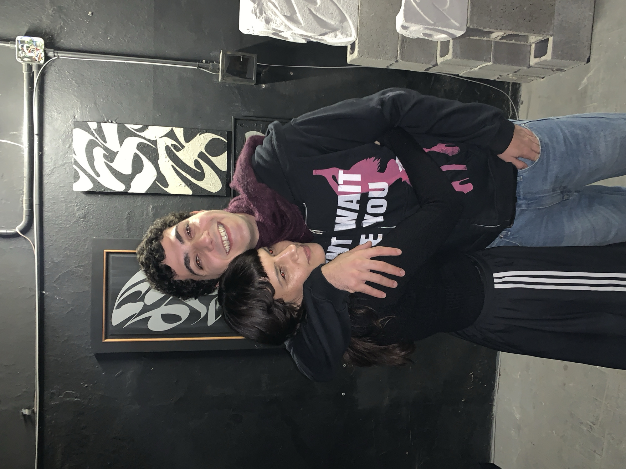 A man and a woman are embracing while lying down. The man is wearing a dark-colored zip-up hoodie with a pink design and the text "DON'T WAIT FOR YOU," blue jeans, and a burgundy hooded sweatshirt underneath. The woman is wearing a black long-sleeved shirt and black pants with white stripes. They are lying in front of a black wall with two framed artworks, one with a black-and-white swirling design, and the other with a silver-and-black design. There is exposed electrical conduit and a junction box on the wall. The floor appears to be concrete. Some white geometric objects are in the upper background. There is no clear indication of the exact location in Buenos Aires, Argentina from the image alone.