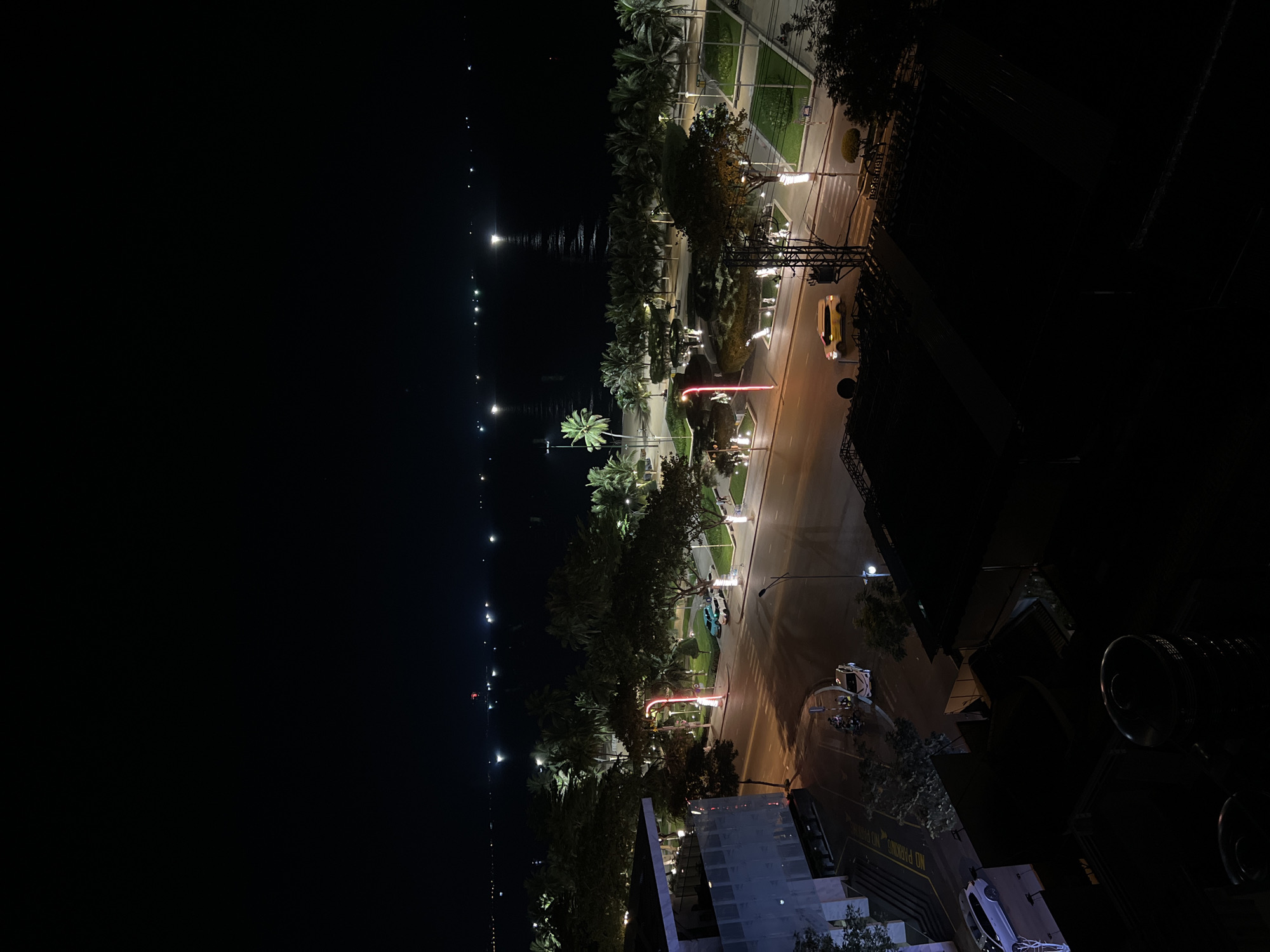 This is an aerial nighttime photograph of a street in Nha Trang, Vietnam, likely near the beach. The scene is dominated by dark skies and the reflections of lights on the water.
In the foreground, the top of dark buildings with corrugated roofs are visible. A tall, cylindrical object, possibly a water tank, is in the lower center. The street below is illuminated by streetlights and the headlights of vehicles. Several cars, including a white sedan and a yellow minivan, are visible on the road. A few motorcycles are parked on the side.
Beyond the street, a paved walkway lined with palm trees and other trees is illuminated by decorative lights. The walkway leads to a dark body of water, presumably the ocean, where distant lights reflect on the surface. The overall impression is one of a tropical city at night, with urban infrastructure bordering a natural seascape.