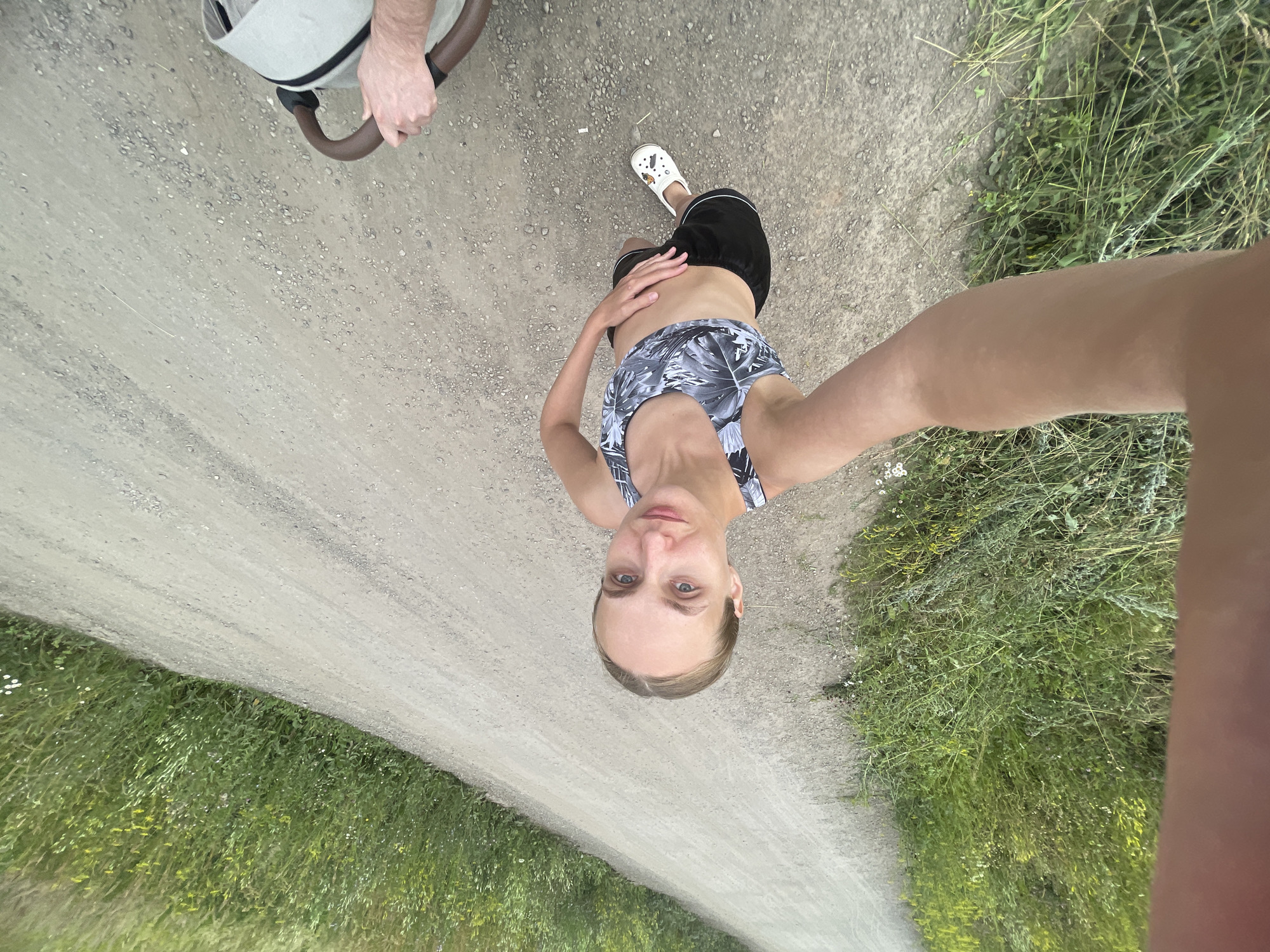 A full-body selfie is taken from a high angle, looking down at a woman standing on a gravel road bordered by tall green grass. The woman is wearing a black and white patterned sports bra and black shorts, with white Crocs on her feet. Her hair is pulled back in a bun, and she is looking at the camera with a slight smile. Her right arm is extended towards the camera, as if she is holding it. To the right of the frame, a person's hands are visible holding the handle of a stroller. The environment appears to be rural, with a long, winding road and lush vegetation on either side. The time of day appears to be daytime, with natural light illuminating the scene. The location is Podshivalovo, Russia.
