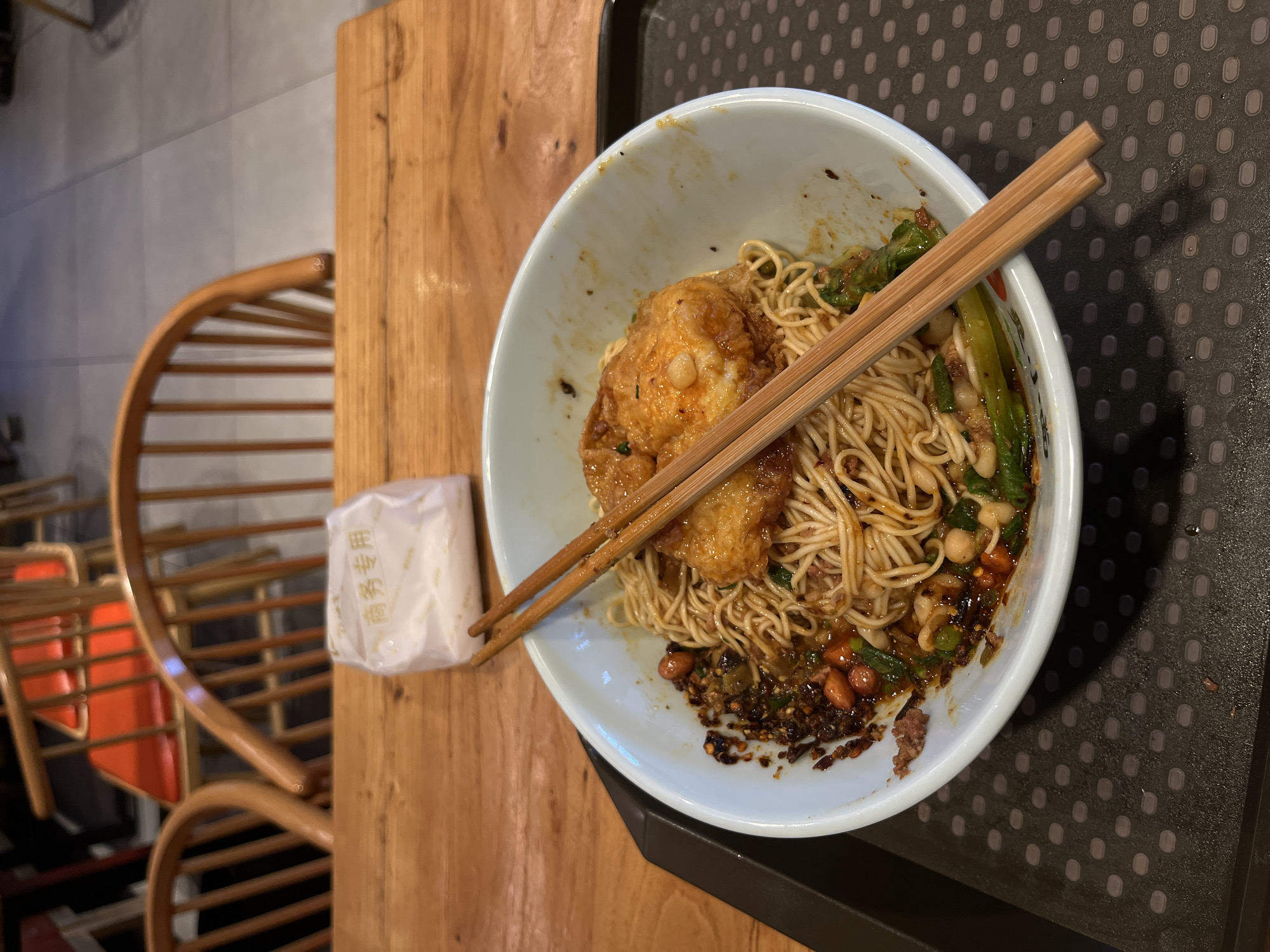 A medium shot, taken indoors, shows a bowl of noodles on a tray, with a packaged item and a chair partially visible. The bowl sits on a dark gray tray with a dotted pattern. Inside the bowl are noodles, a fried item, green vegetables, and a dark sauce with peanuts. A pair of wooden chopsticks rests across the top of the bowl. To the left of the bowl, on a wooden surface, is a small, white, wrapped item with Chinese characters printed on it. A wooden chair with a circular back is visible in the background. The environment suggests a dining establishment, possibly located in the Yuzhong District of China, given the likely regional cuisine. There are no people visible in the image.