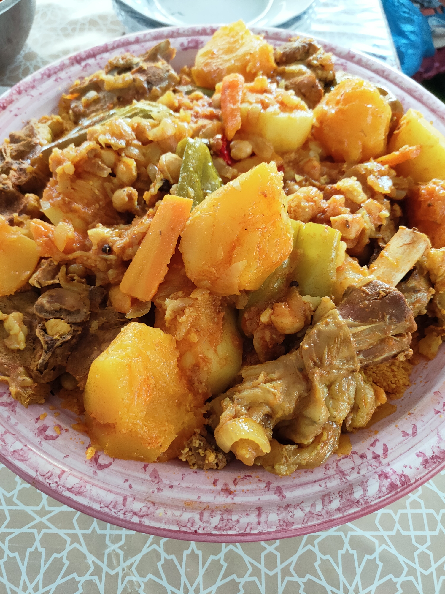 A close-up, overhead shot captures a large, pink-rimmed ceramic plate filled with a hearty stew. The stew is a rich, orange-brown color, indicating a tomato-based sauce, and is generously filled with large chunks of golden-yellow potatoes, sliced carrots, and pieces of meat, some of which appear to be lamb or mutton with bone fragments visible. Green bell pepper pieces are also interspersed throughout the dish, along with what seem to be chickpeas. The food is piled high, suggesting a generous serving. The plate rests on a light-colored tablecloth with a repeating geometric pattern in a lighter beige. The scene appears to be an indoor setting, likely a dining area, though the broader environment is not visible. The image quality is good, allowing for clear identification of the food items and their textures. The photograph was taken in the city of Ariana, Tunisia.