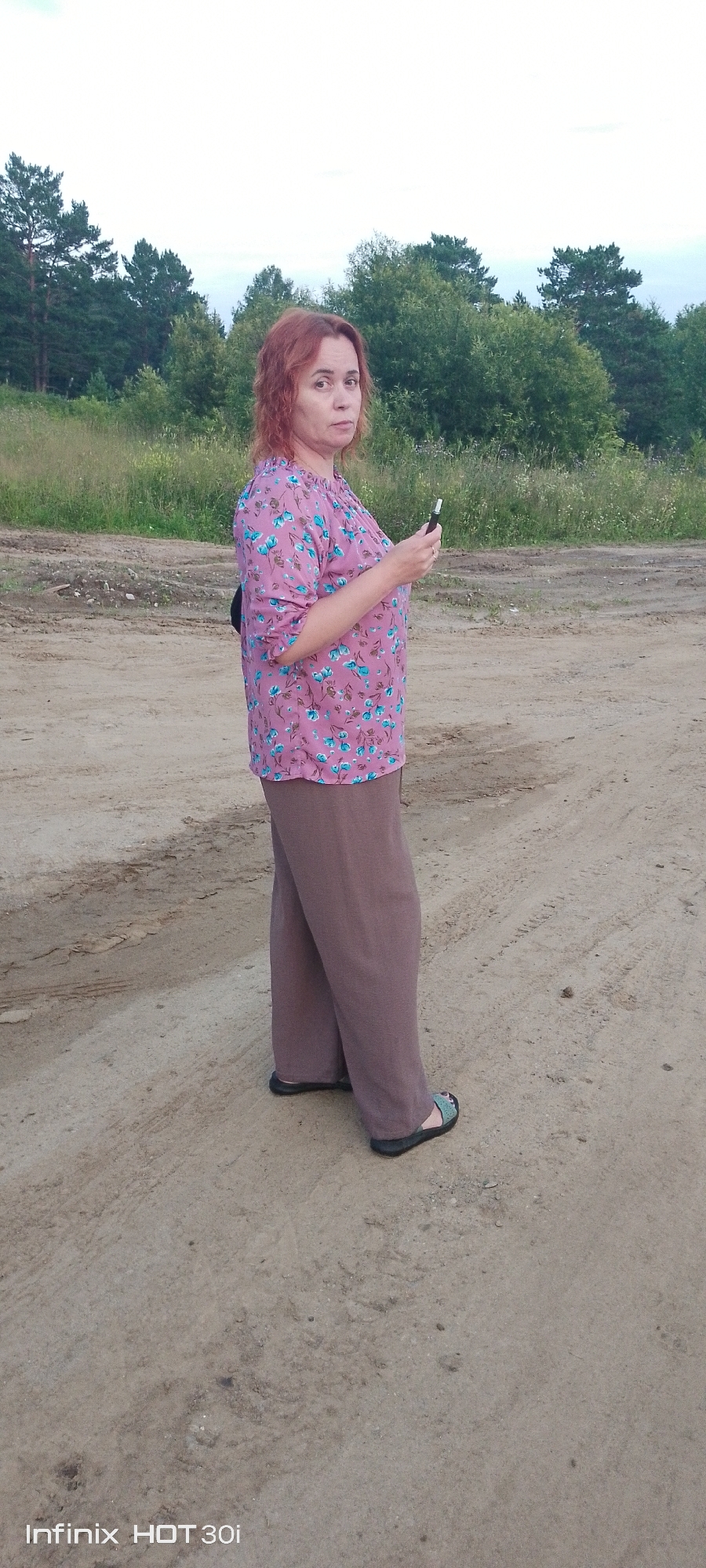 A woman with reddish-brown, shoulder-length hair stands in a dirt clearing, looking towards the camera with a neutral expression. She is wearing a pink blouse with a floral pattern of blue flowers and green leaves, and loose-fitting, taupe-colored pants. On her feet are dark sandals with a light-colored patterned strap. In her right hand, she holds a dark, slender object that appears to be a vape pen or e-cigarette. She is positioned in the foreground, with a dirt path and tire tracks visible on the ground around her. In the background, a line of trees, mostly green with some coniferous varieties, stretches across the horizon under a bright, overcast sky. The overall setting suggests an outdoor, possibly rural or semi-rural area, consistent with the outskirts of Kedrovy, Russia.