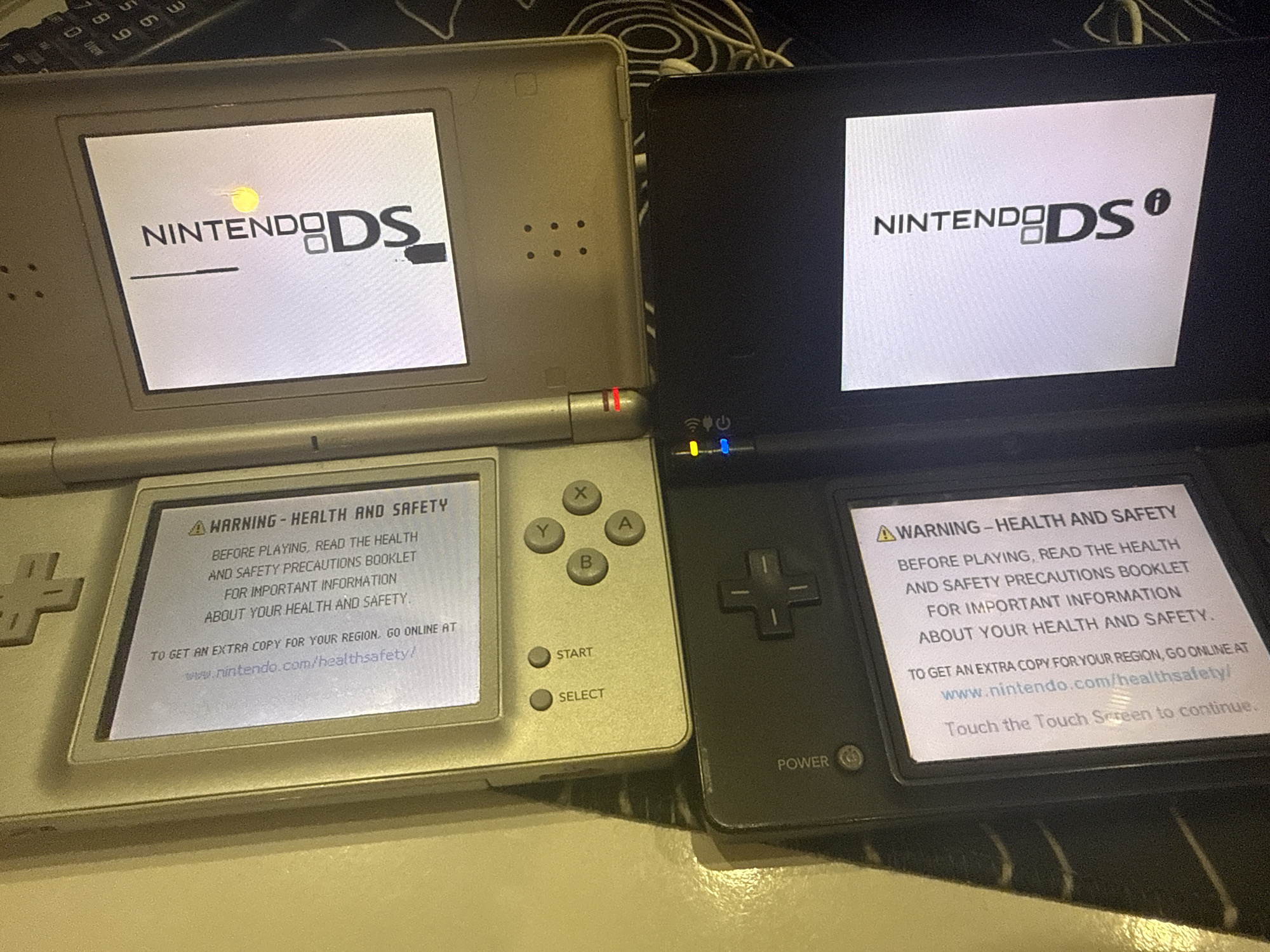 The image shows two Nintendo DS handheld gaming consoles. The top console is silver, and the bottom console is black. Both consoles are displaying a warning message related to health and safety. The silver console's screen shows a warning about reading the health and safety precautions booklet before playing. The bottom console's screen also shows a similar health and safety warning. There are visible buttons and controls on both consoles. The environment appears to be indoors, likely a domestic setting, but there are no discernible landmarks or features that would allow for identification of a specific location within Burgas, Bulgaria. The lighting suggests the photograph was taken in an environment with artificial light.